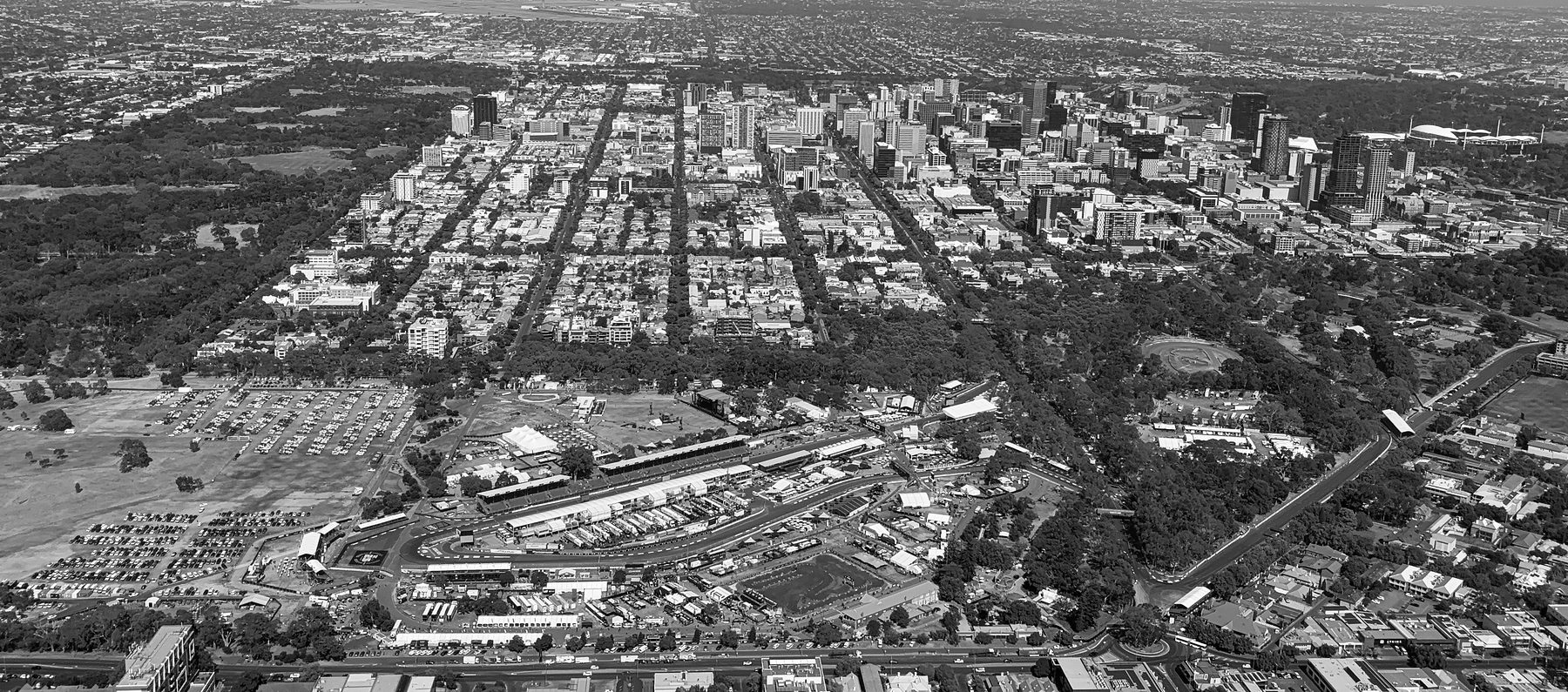An aerial view over a city’s parklands: a temporary pit straight, grandstands and paddock tents in the foreground, rows of parked cars to the left, and the dense grid of the city and its towers behind.