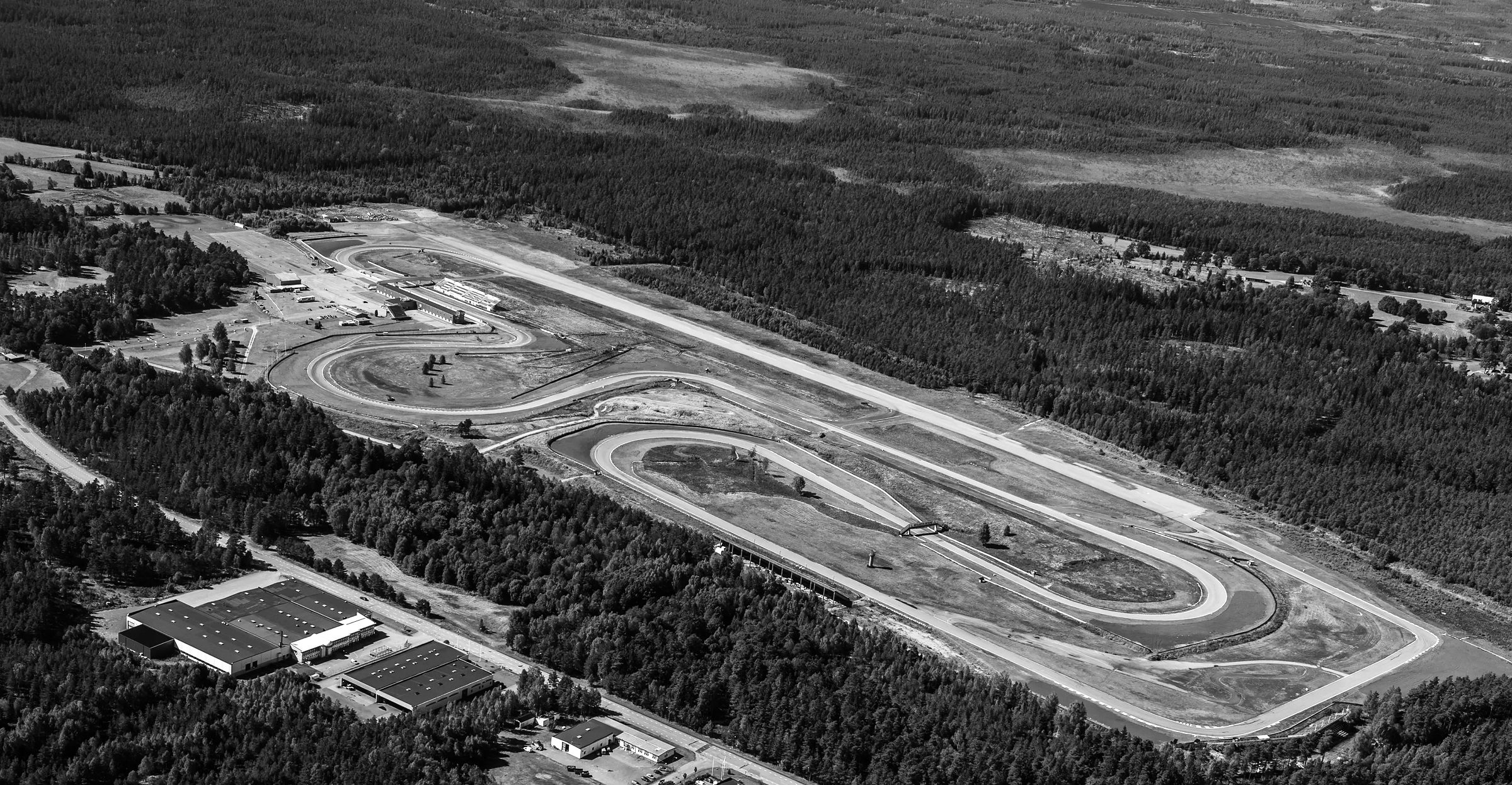 An aerial view of a racing circuit laid out in a pine forest: a long straight runway beside loops of track, pit buildings at the far end, open bog and forest stretching to the horizon.