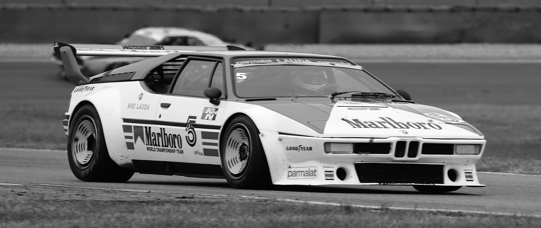 A low, wedge-shaped racing coupé with a tall rear wing and No. 5 on its flank corners on a circuit, the driver’s helmet visible through the windscreen; another car runs on the track behind.