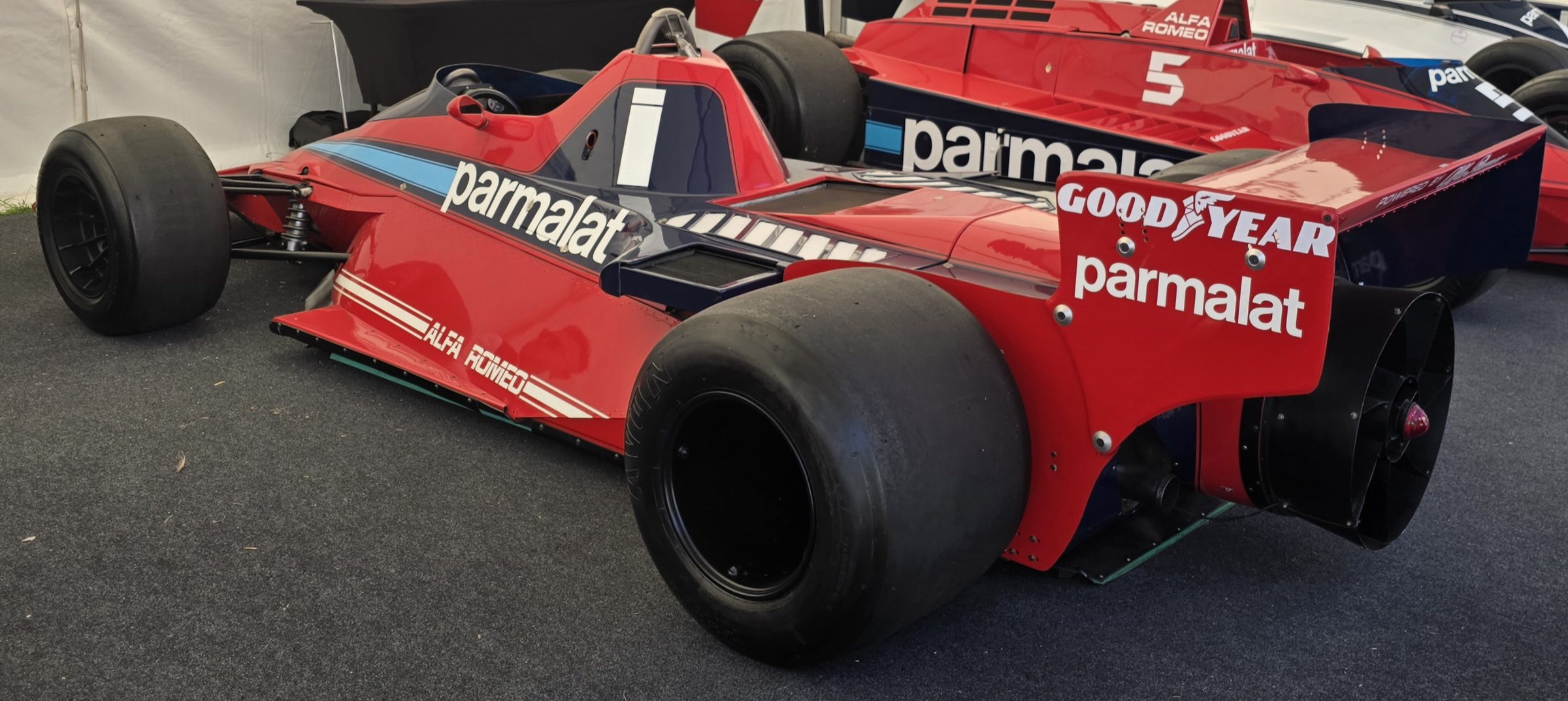 In colour: a red Formula One car seen from the rear three-quarter, Parmalat on its side and rear wing and a large circular fan at its tail; a second red car with No. 5 stands behind it in a white tent.