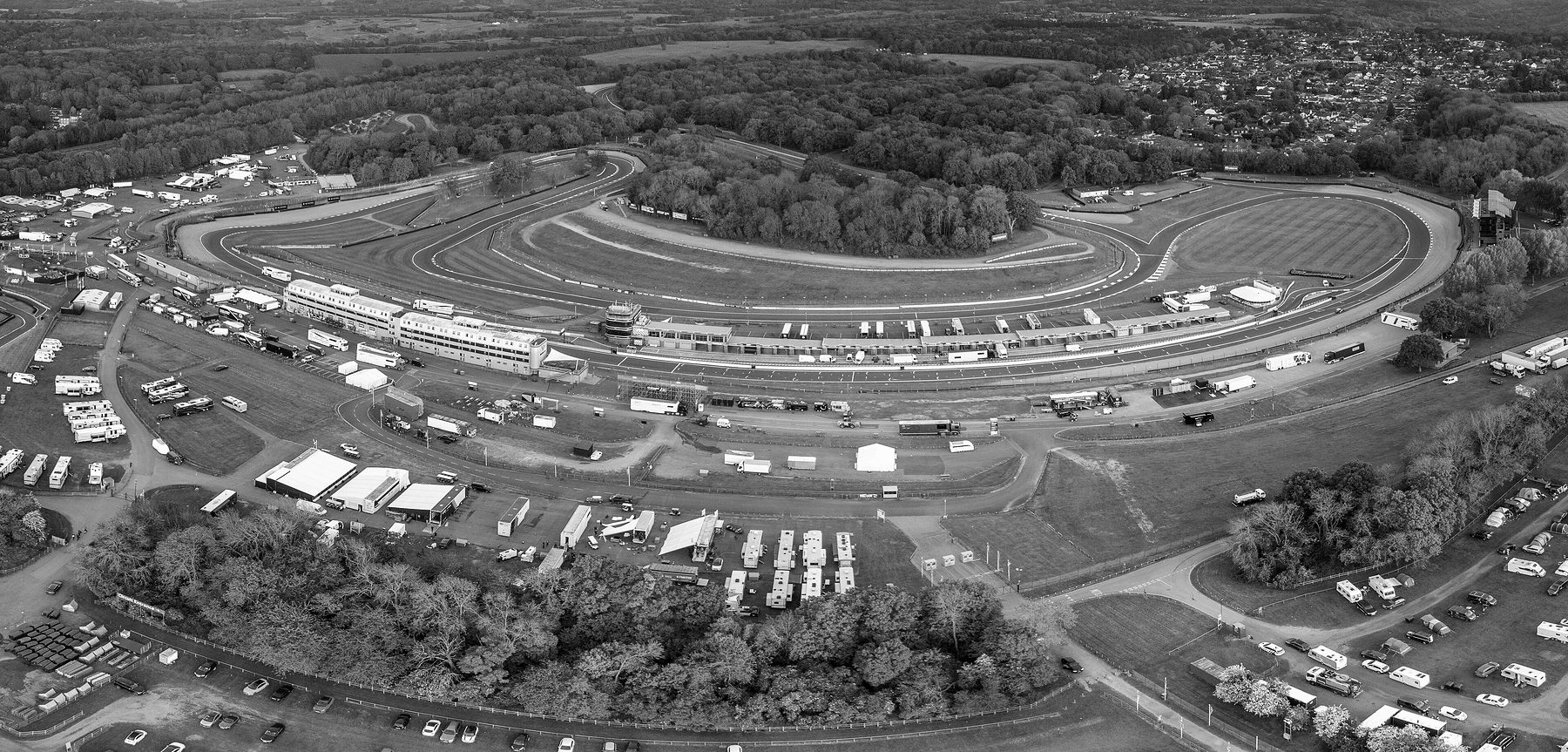 An aerial view of a racing circuit in wooded country: the track curving round a grassy infield, pit buildings and a long garage block along the straight, transporters and motorhomes in the paddock in front.