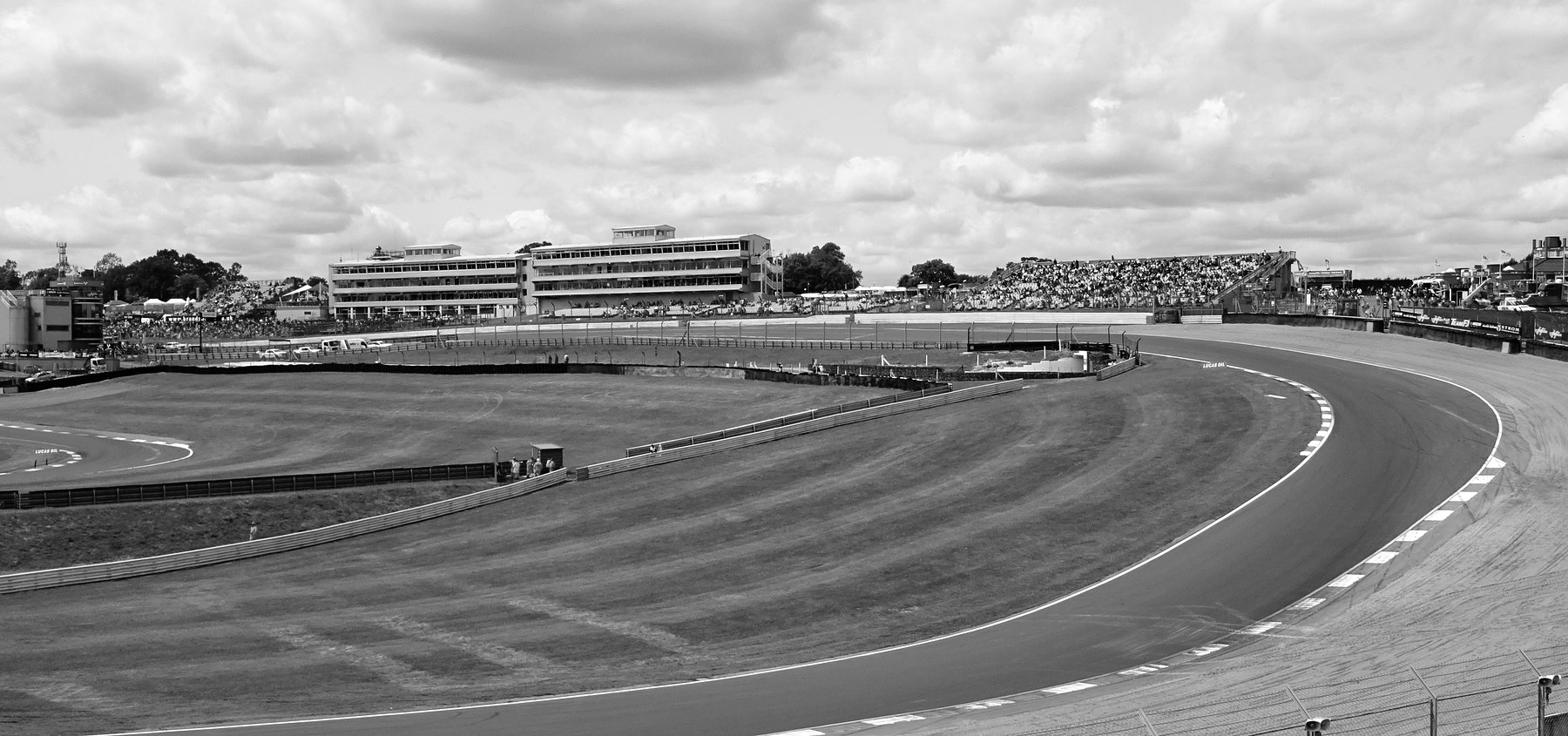 A downhill right-hand bend with striped kerbs and a gravel trap on its outside, a mown grass infield with marshals at a post, and a modern pit building and a packed grandstand under a cloudy sky.