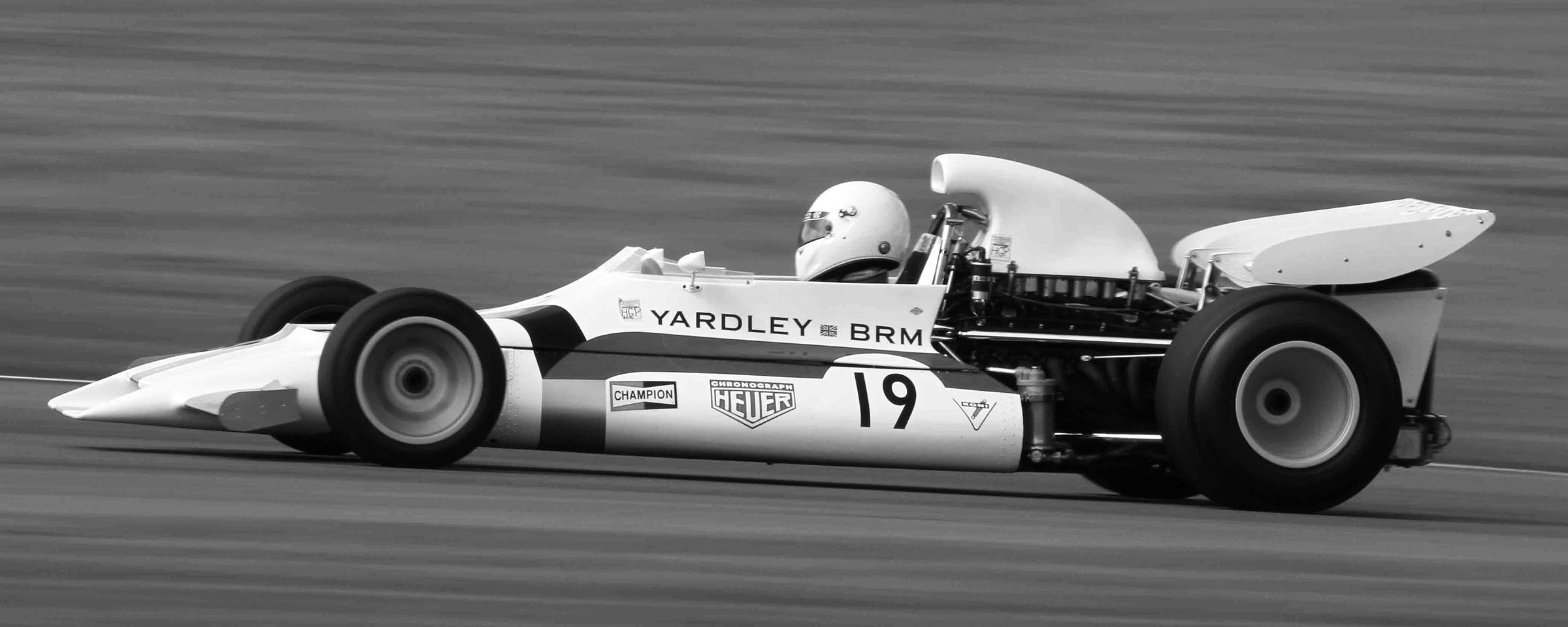 A slim, wedge-nosed single-seater with a tall air intake behind the driver’s white helmet and No. 19 on its side, panned at speed; grass and the edge of the track streak past behind it.