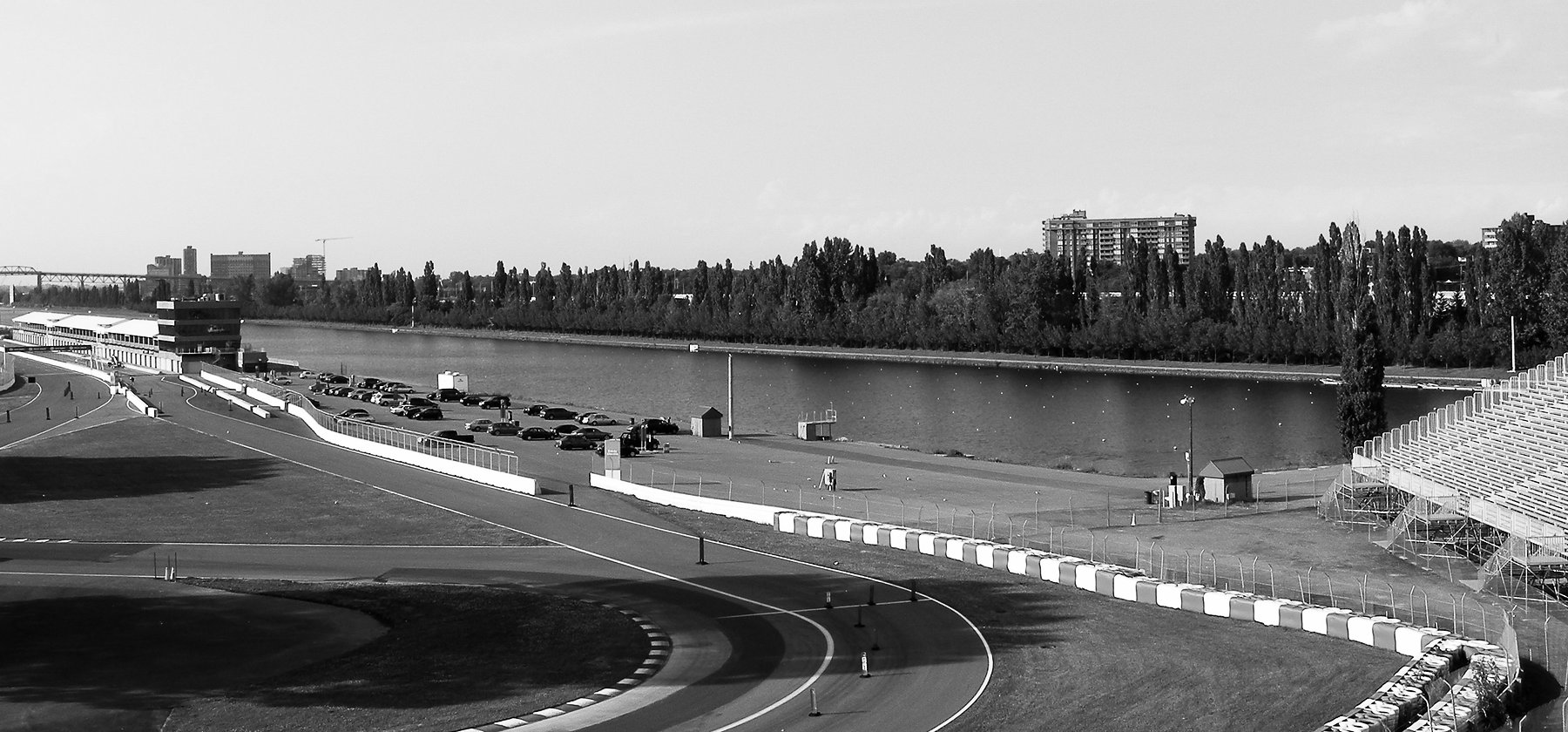 A circuit on an island: a sweeping bend with striped kerbs and a long run of striped tyre barriers in the foreground, an empty paddock apron and a temporary grandstand, a long rowing basin lined with poplars, and city towers and a bridge on the far left.