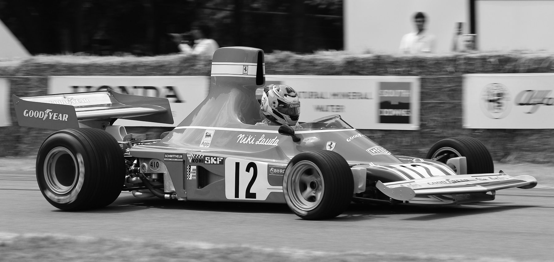 Ein flacher Monoposto mit der Nummer 12, einem hohen Lufteinlass hinter dem Helm des Fahrers und einem breiten Heckflügel fährt an Strohballen und Werbetafeln vorbei; hinter den Ballen stehen einige Zuschauer und ein Fotograf.
