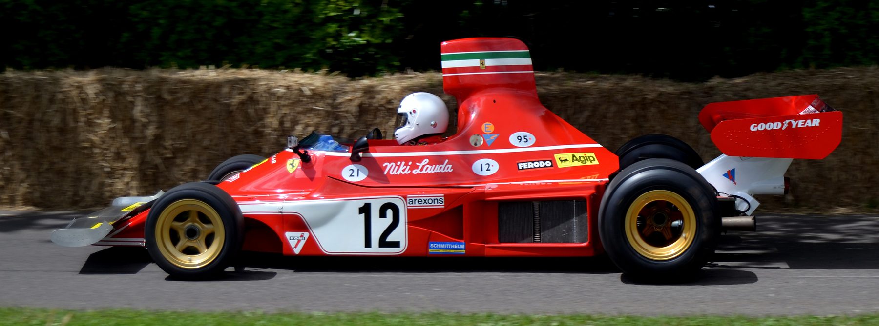 In colour: a red Ferrari single-seater with gold wheels, No. 12 on a white panel and a tall airbox striped green, white and red, seen in profile against straw bales and dark trees; the name Niki Lauda is painted on its flank, and the driver wears a white helmet.