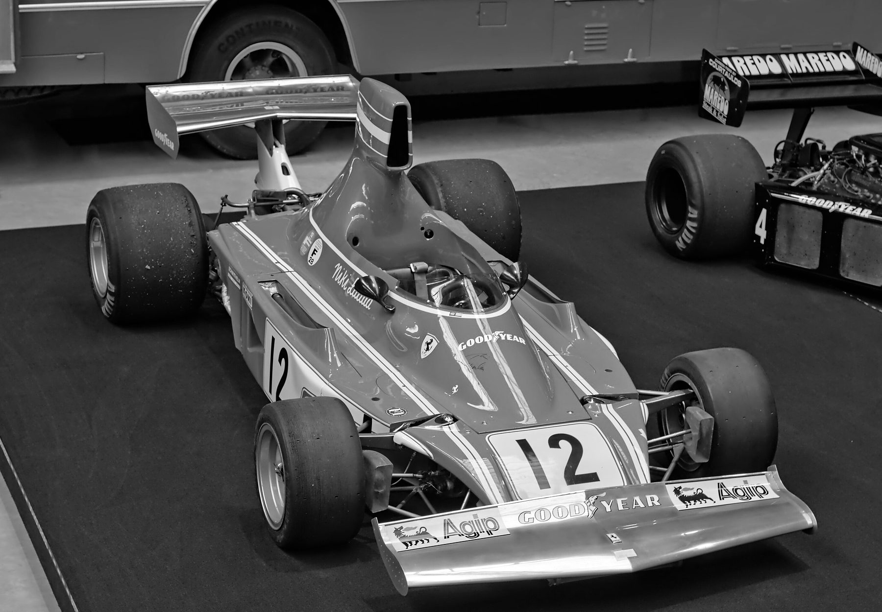 A Formula One car with a tall airbox, No. 12 on its nose and side and a wide front wing stands on a dark carpet in a museum hall; a team transporter is behind it, and the rear wing and wheels of another car show at the right.