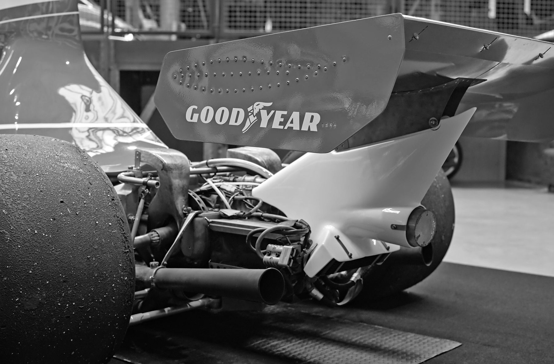 Close view of the back of a Formula One car: a large rear wing with Goodyear lettering above an exposed gearbox, wiring and a stubby exhaust pipe, with a scuffed rear tyre filling the left of the frame.