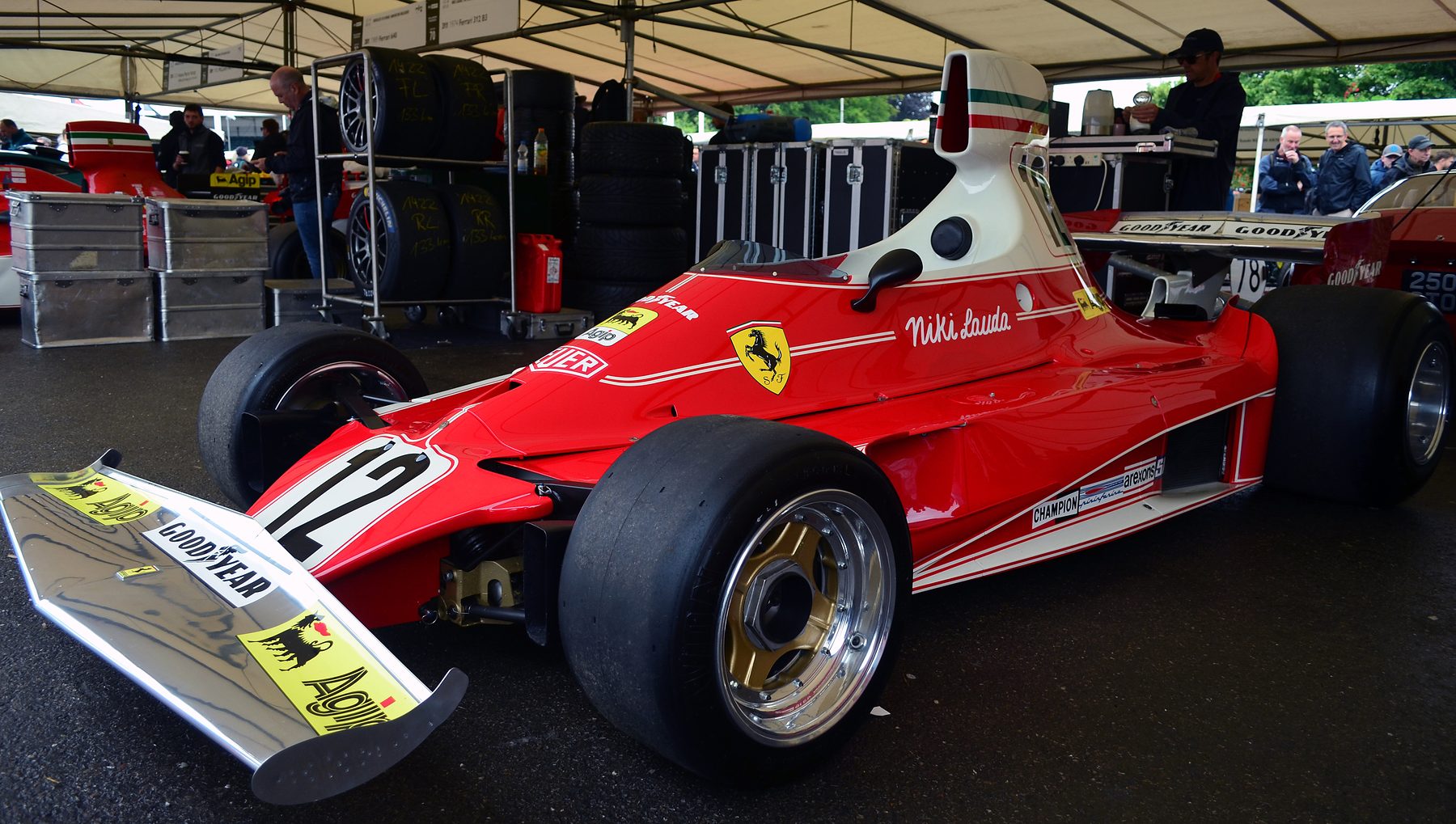 In colour: a red and white Ferrari single-seater with a tall white airbox, No. 12 on its nose and the name Niki Lauda on its flank, parked under a paddock awning, its polished front wing in the foreground; tyre racks, flight cases and onlookers behind.