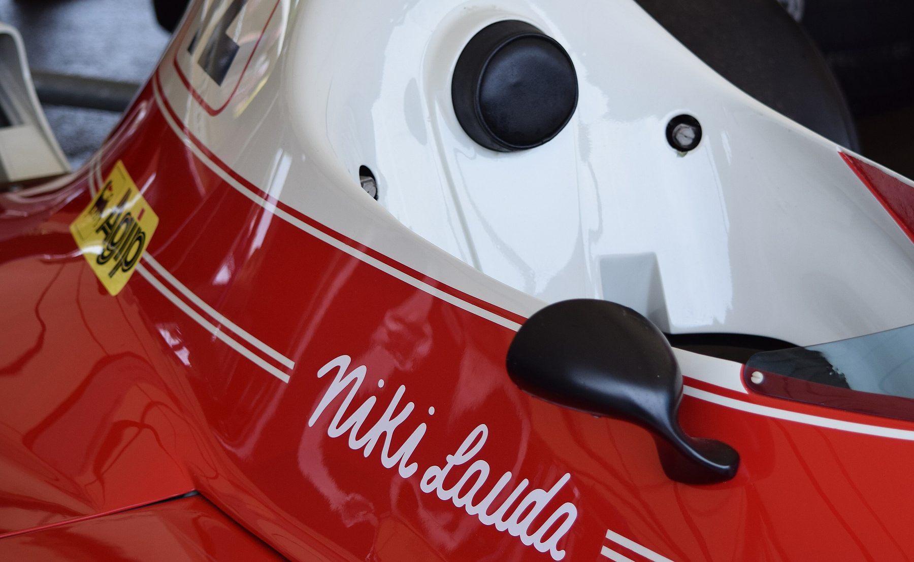 In Farbe: Nahansicht der Flanke eines roten Ferrari unter einer weißen Cockpitumrandung, mit „Niki Lauda“ in weißer Schreibschrift, einem schwarzen Spiegel und Zierlinien.