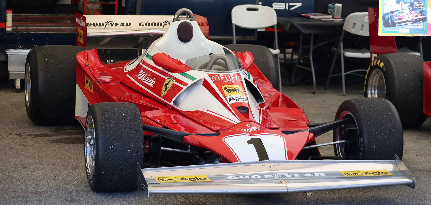 In colour: head-on view of a red and white Ferrari single-seater with a low cockpit and No. 1 on its nose, parked in front of a team transporter, folding chairs and a table.