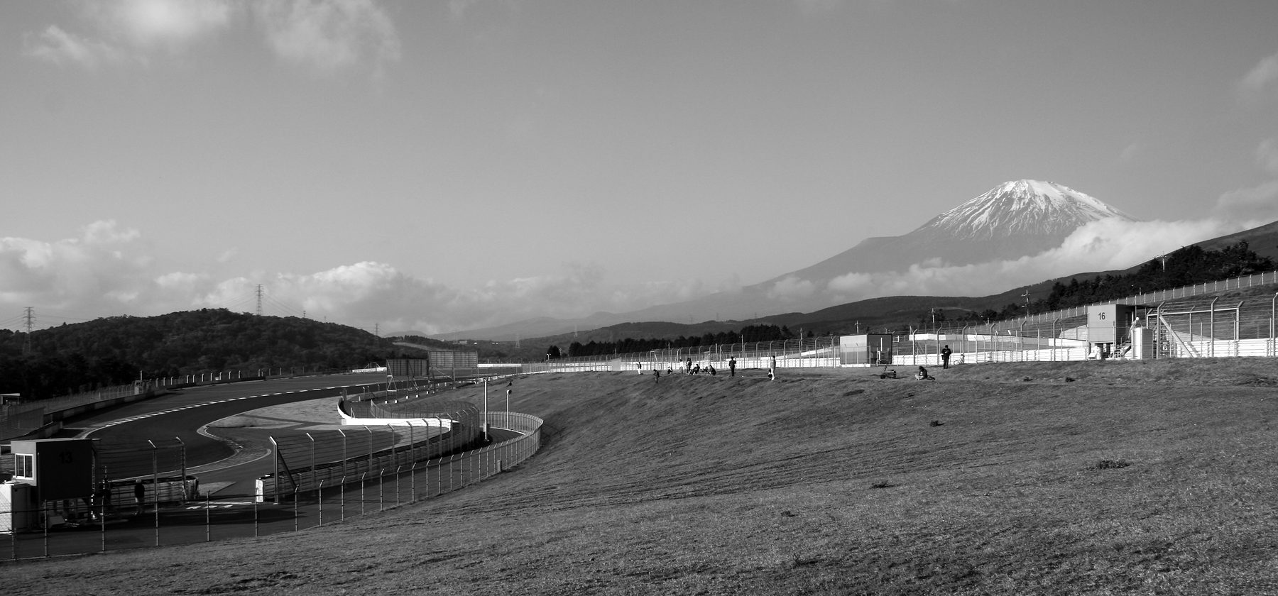 A grassy spectator bank above a racing circuit, the track curving away on the left behind fences, a few people on the bank, and the snow-capped cone of Mount Fuji rising above a band of cloud.