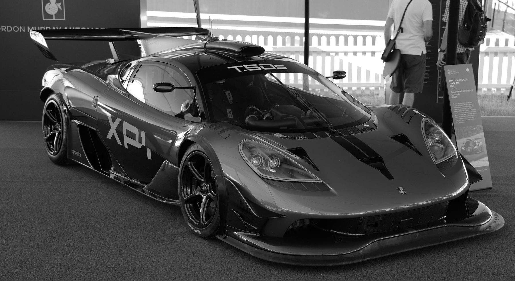 A low mid-engined track car with a large rear wing, a roof scoop and XP1 on its flank, on a carpeted display stand; two visitors and an information board stand behind it.