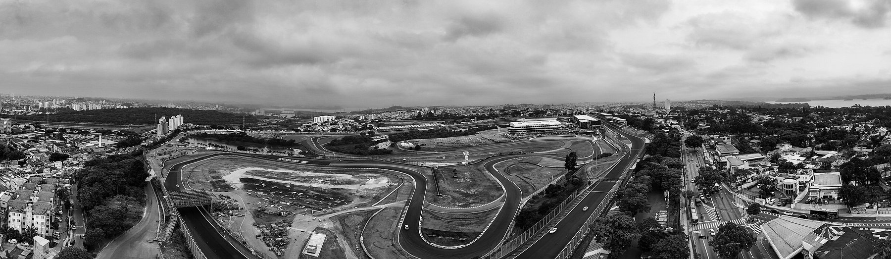 A wide aerial panorama of a racing circuit among city suburbs: the track winding through a grassy infield, a long pit building and grandstands, cleared ground with stacked scaffolding on the left, and streets and houses stretching to a lake under heavy cloud.