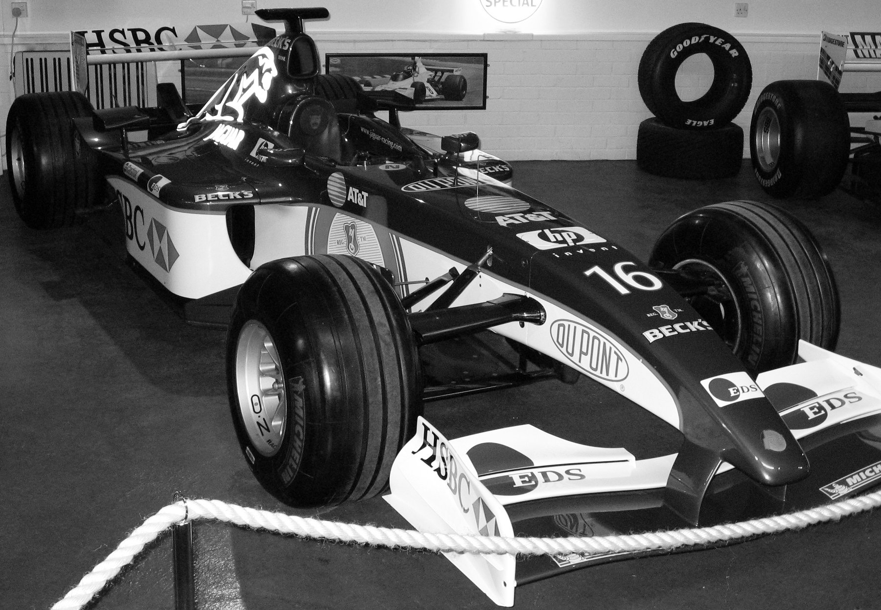 A Formula One car of the early 2000s with No. 16 on its nose and a leaping cat on the engine cover, on a museum floor behind a rope barrier; spare tyres and photographs on the wall behind.