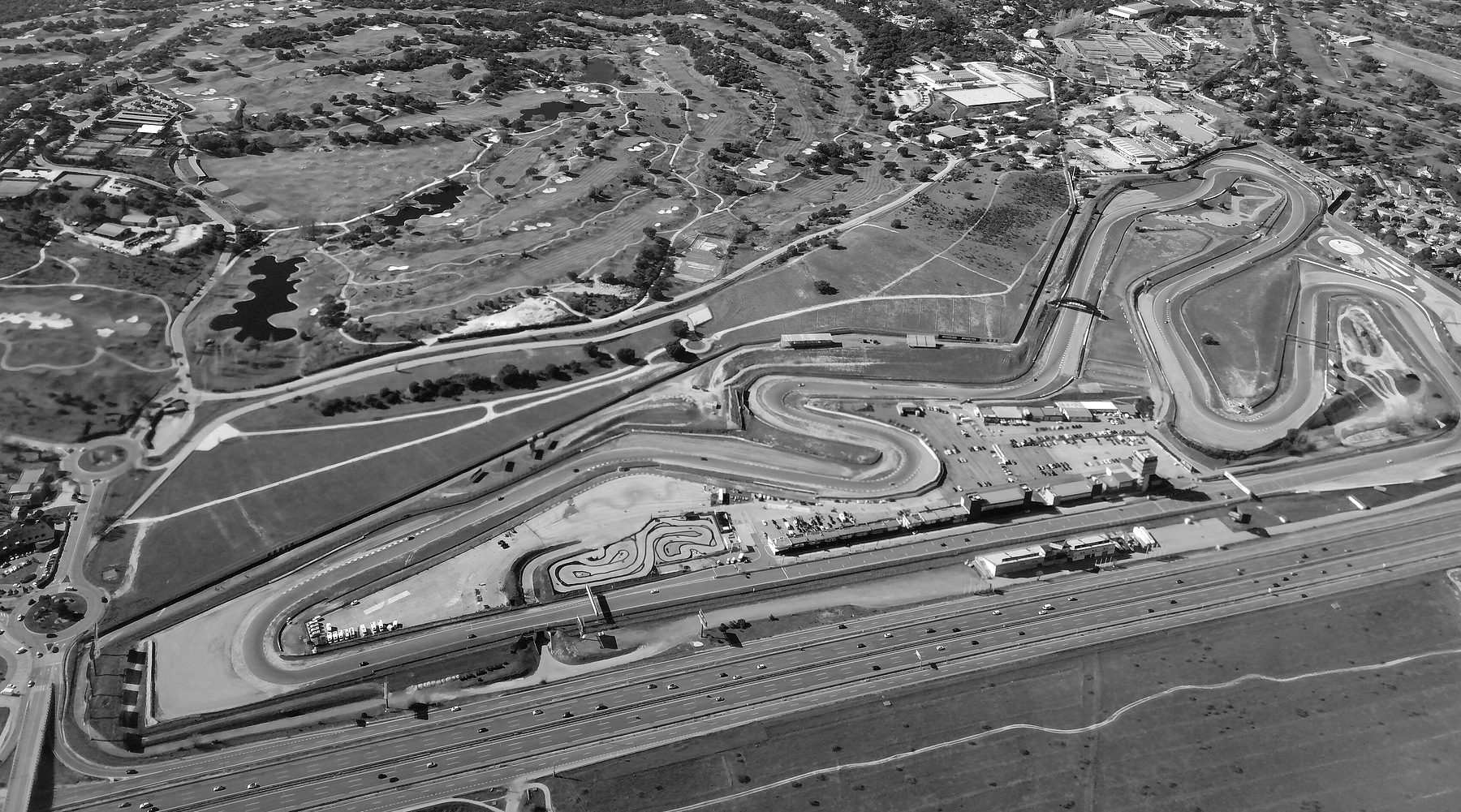 An aerial view of a twisting racing circuit on dry open ground beside a wide motorway, a car park and pit buildings in the middle, a golf course and suburbs beyond.