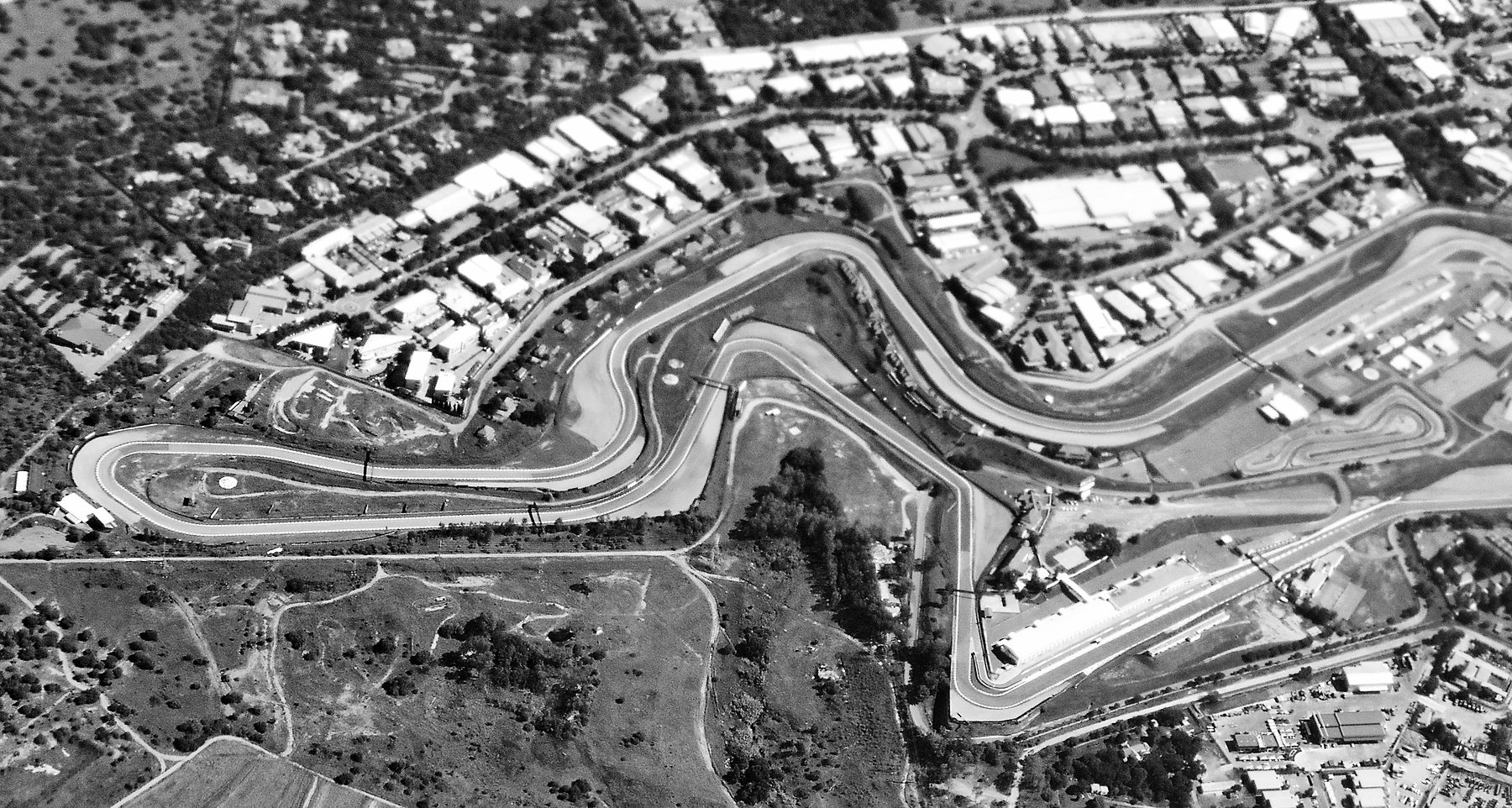 An aerial view of a racing circuit snaking through scrubland, with sandy run-off areas, a pit straight and paddock at the right, a kart track, and blocks of industrial sheds and houses around it.