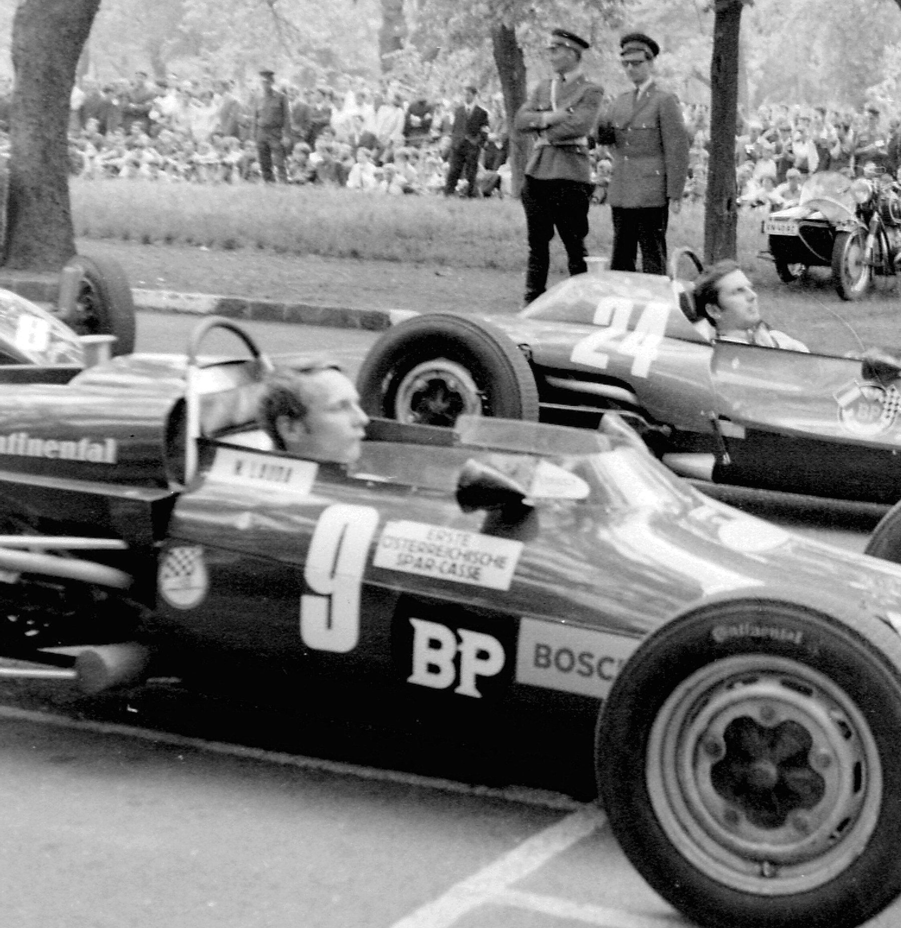 A young bare-headed driver sits low in a single-seater, No. 9, its flank covered in sponsors’ lettering; beside it a second car, No. 24, its driver looking up; behind them two uniformed policemen stand, one with his arms folded, before a large crowd under the trees.