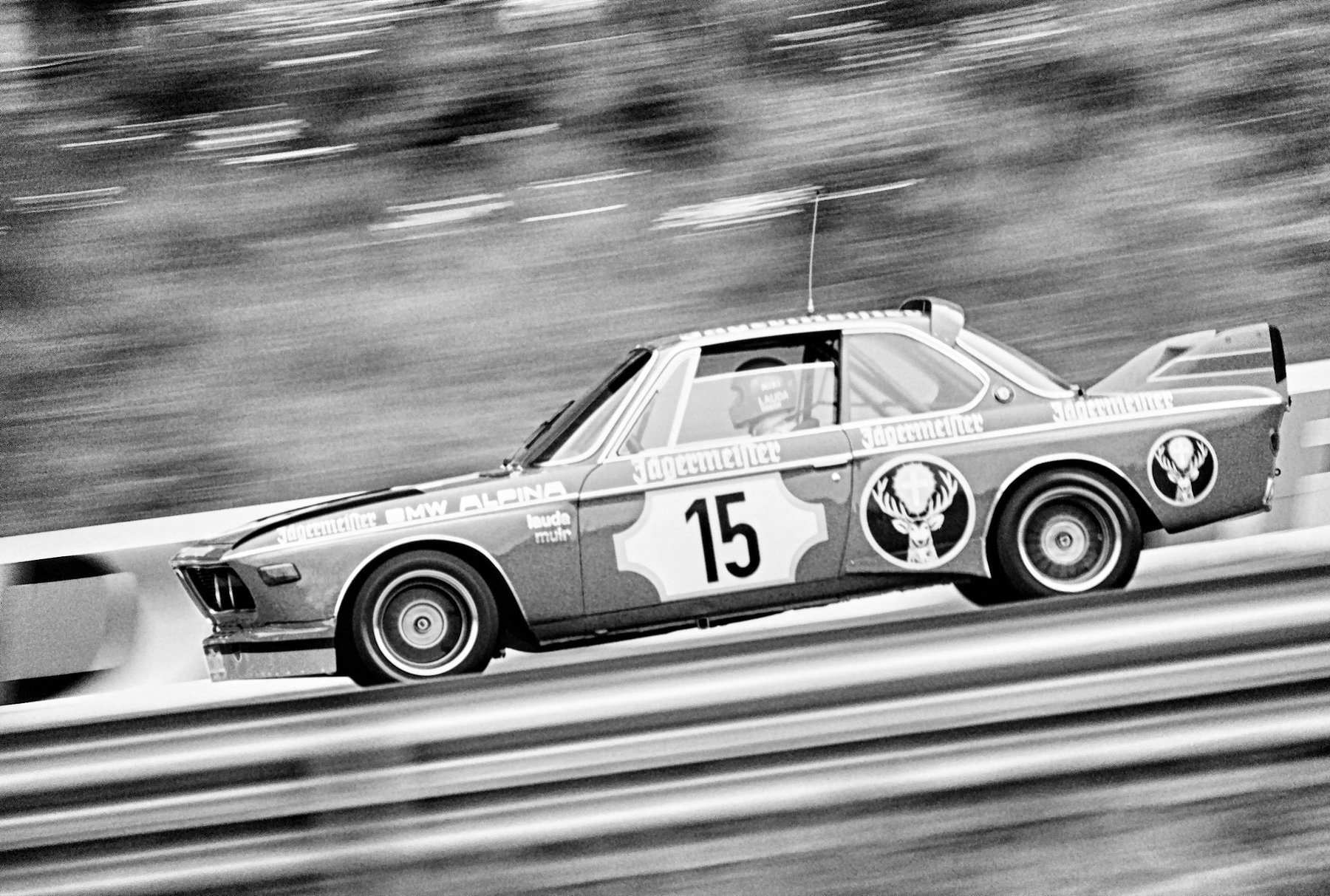 Panned side view of a saloon racing car, No. 15, at speed beside a steel barrier, the trees behind streaked into blur; a large rear wing, a stag emblem on the door and the name LAUDA on the driver’s helmet through the side window.