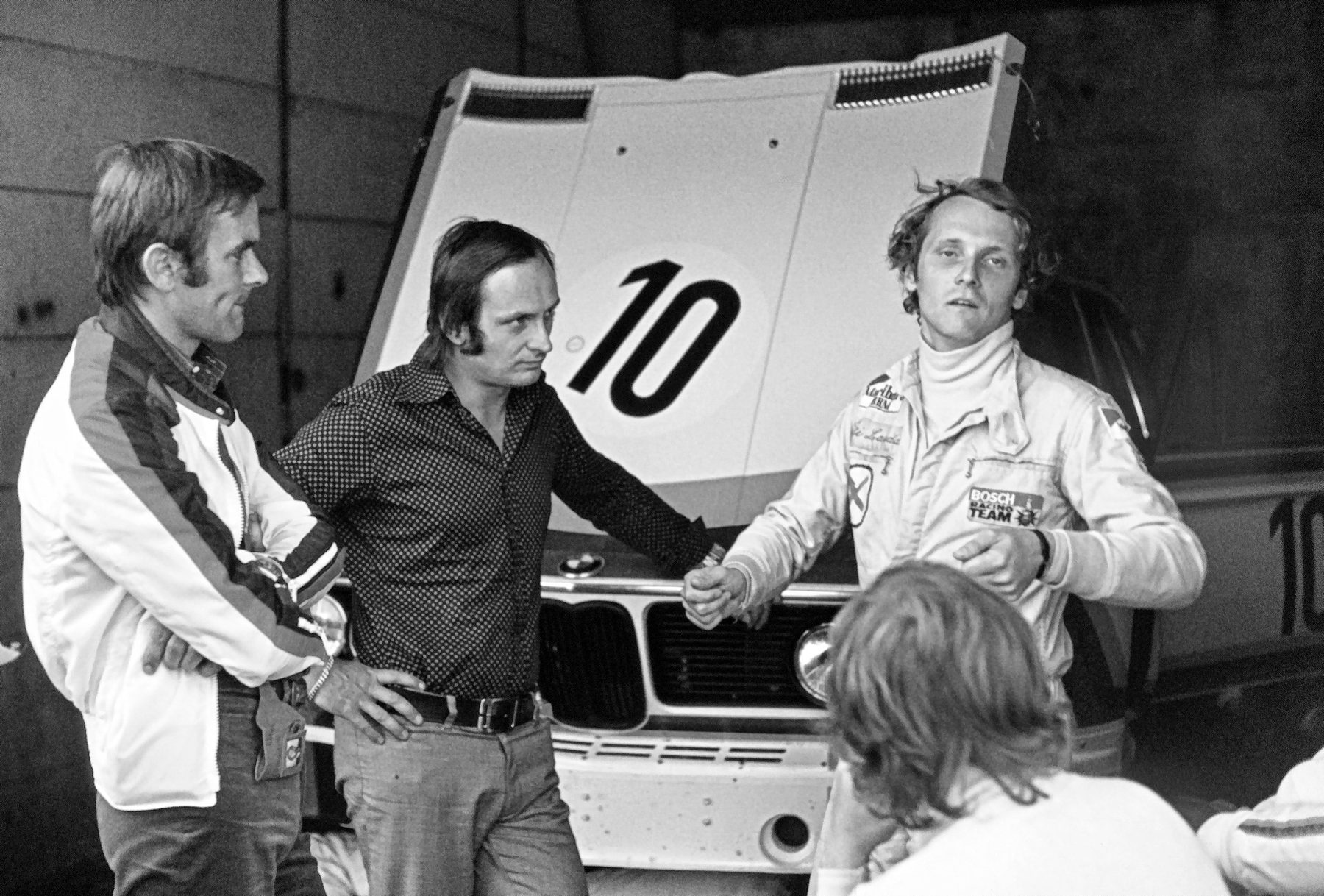 A young man with tousled hair, in white racing overalls and a roll-neck, talks with his hands in front of the raised bonnet of a saloon car numbered 10; two men stand listening at the left and the back of a head fills the lower right.