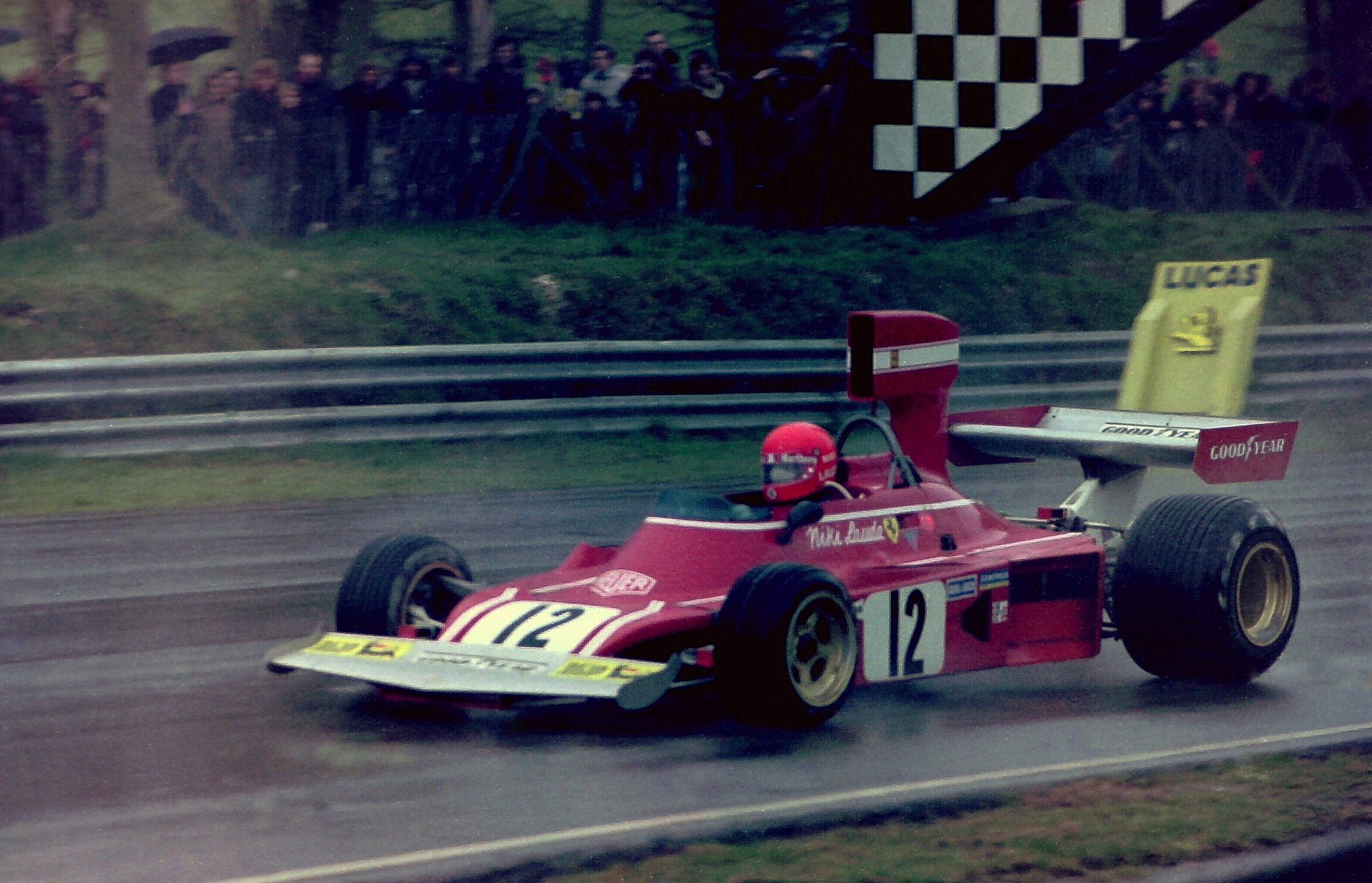 In colour: a red and white Formula One car, No. 12, on a streaming wet track, its driver in a red helmet; spectators with umbrellas and a chequered gantry behind the barrier.