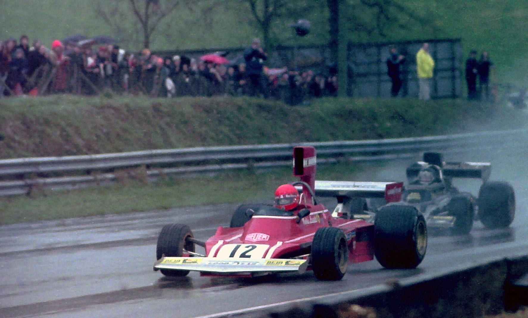 In colour: a red Formula One car, No. 12, its driver in a red helmet, leads a black car through spray on a wet track; spectators under umbrellas line the bank behind.