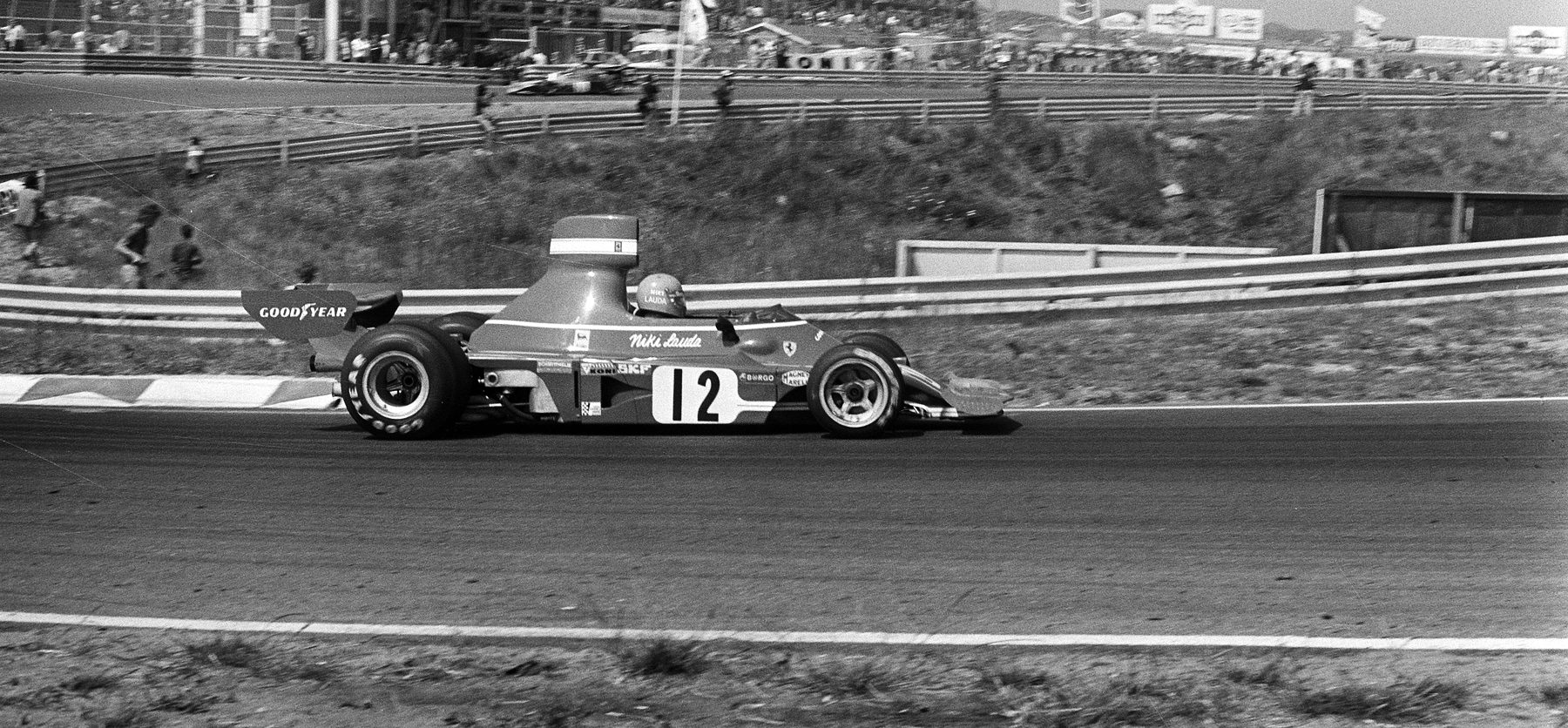Side view of a Formula One car, No. 12, the name Niki Lauda in script on its flank and on the driver’s helmet, on a curving track; behind it a barrier, a grassy bank with a few spectators, and another car on the track above.
