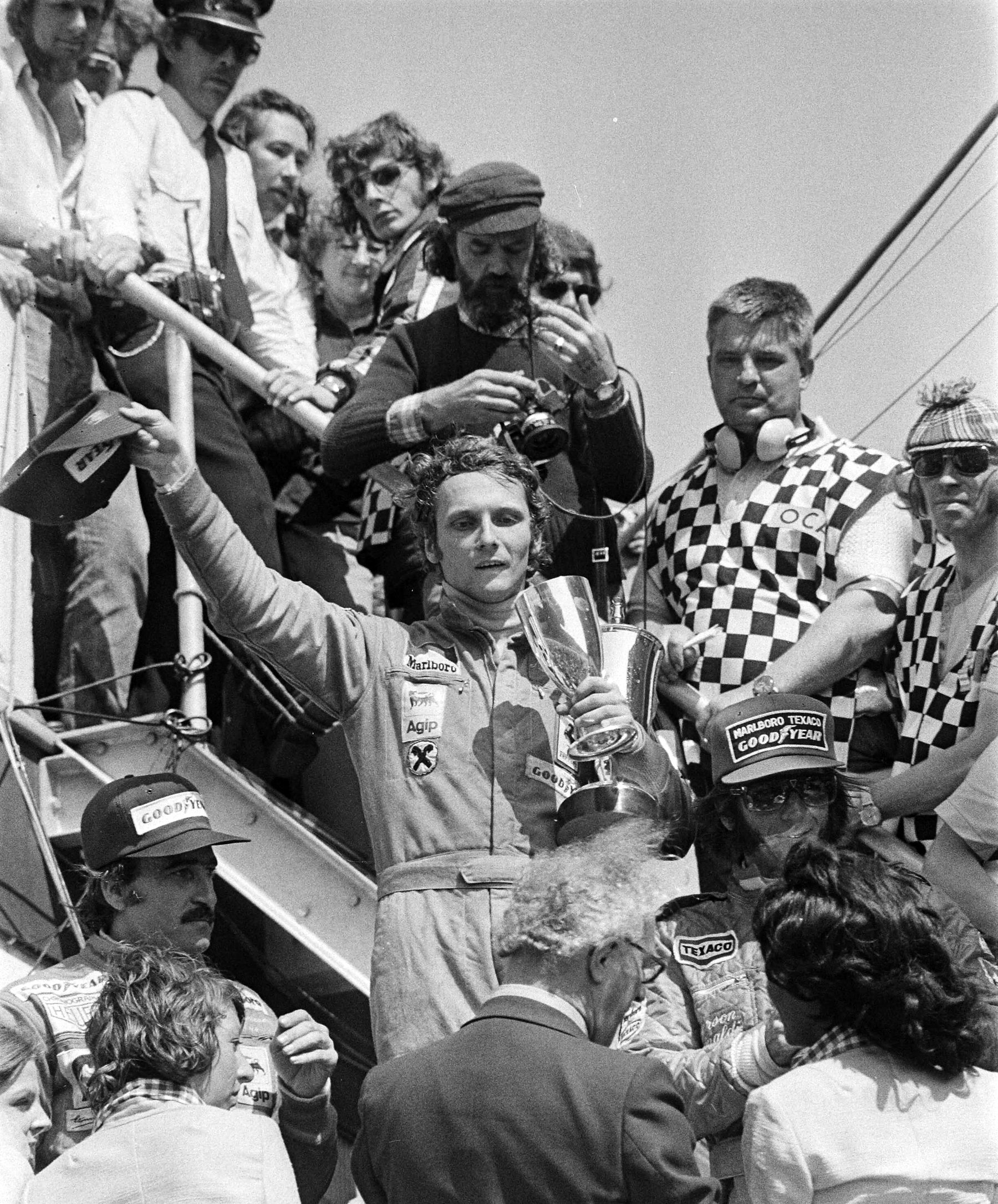 A curly-haired young man in racing overalls raises a cap in one hand and holds a large cup in the other on a crowded podium stair; photographers and men in chequered waistcoats press round him, and a driver in a cap stands at the lower left.