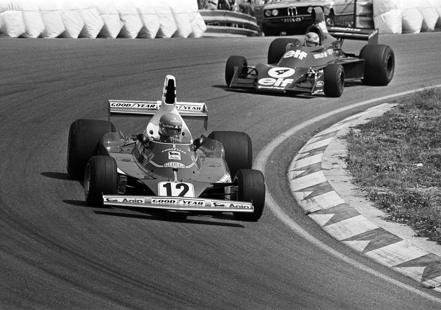 Head-on, a single-seater numbered 12, with a tall airbox behind the driver’s helmet, leads a second car numbered 4 round a bend beside a painted kerb; white sandbags and two parked saloon cars stand behind.
