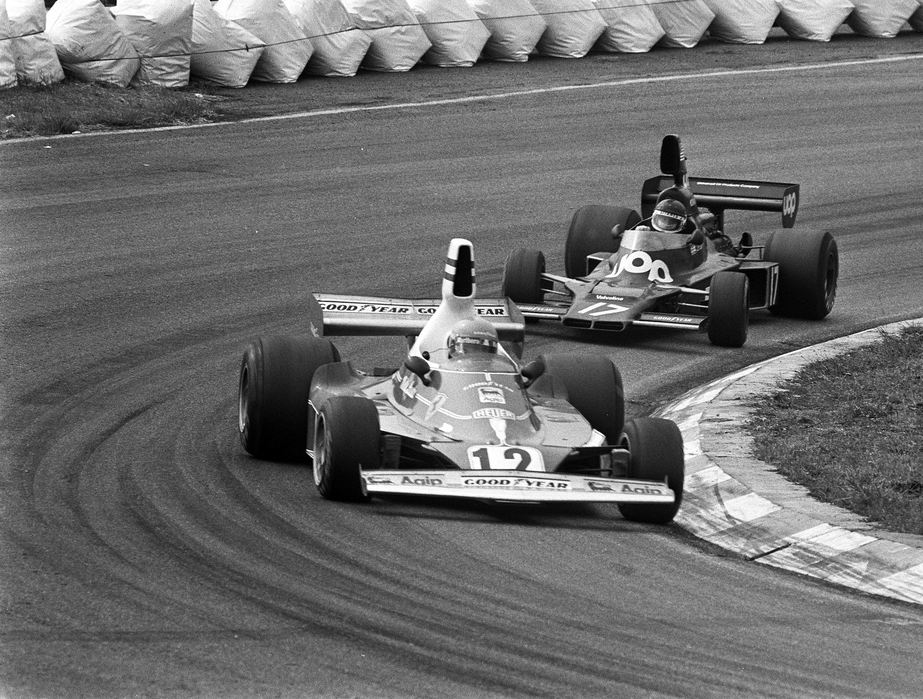 A Formula One car, No. 12, leads a dark car, No. 17, through a curve beside a striped kerb; a row of white sandbags lines the far side of the track.