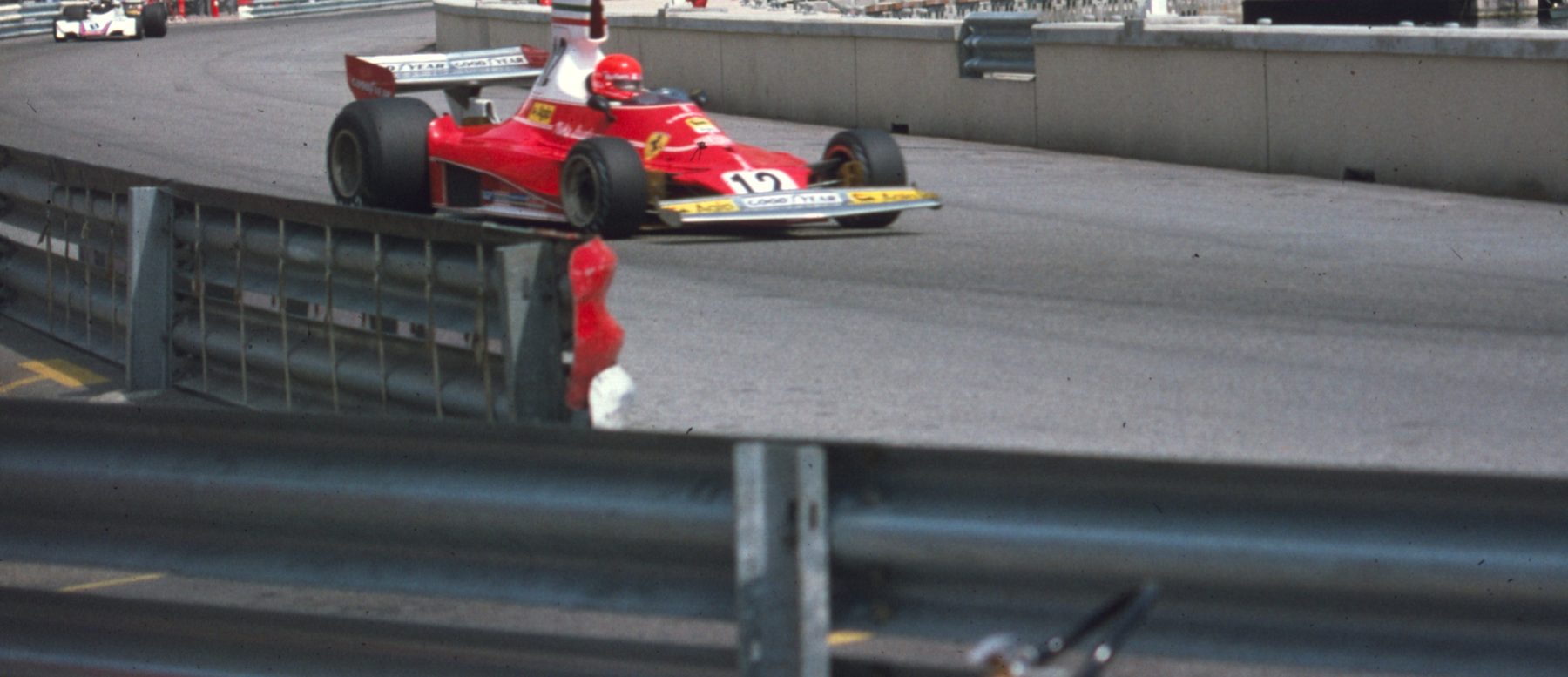In colour: a red and white single-seater numbered 12, its driver in a red helmet, comes out of a corner beside a concrete wall, with marshals and a parked car beyond and a steel barrier in the foreground.