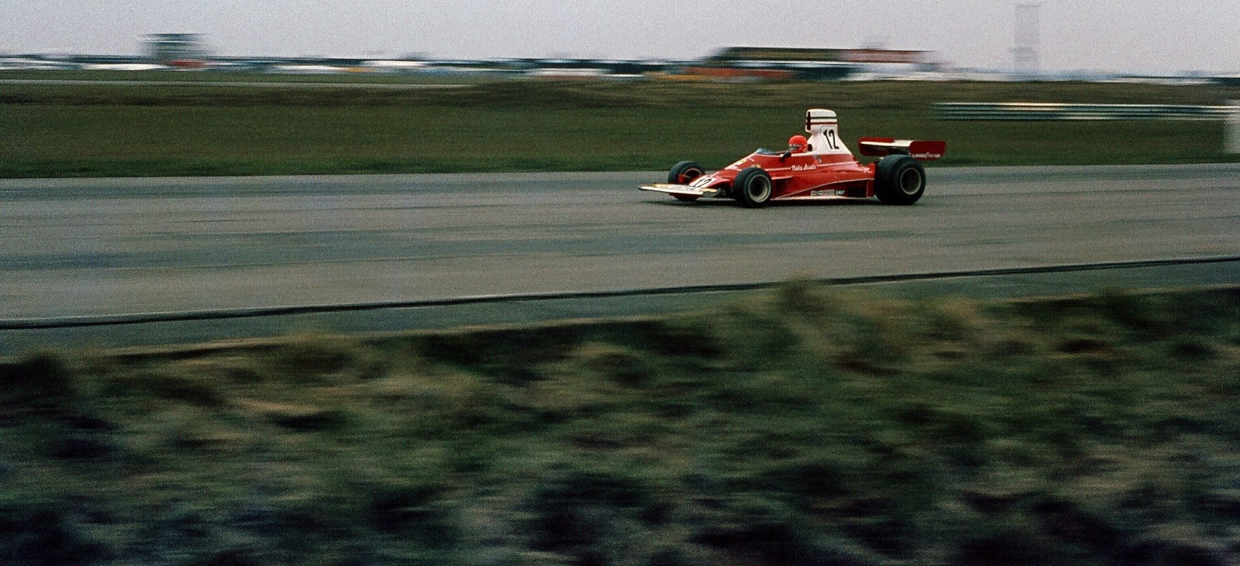 In colour: a red and white single-seater numbered 12 crosses a flat airfield circuit at speed, the grass, the distant stands and the verge in the foreground all streaked by the pan.