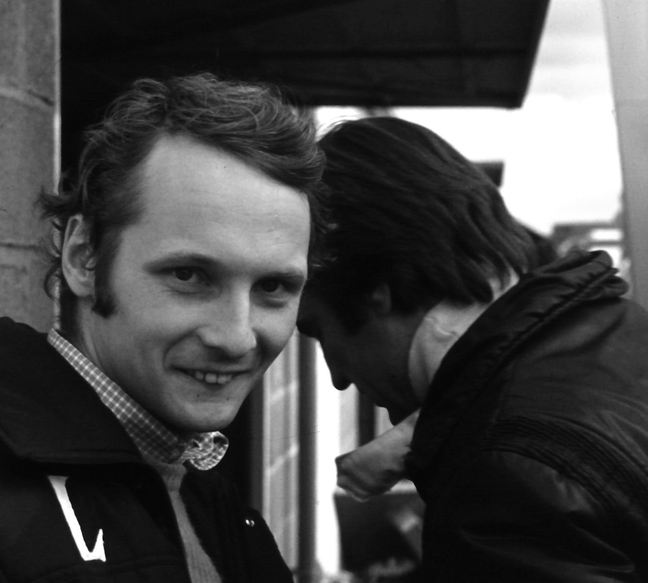A smiling young man with swept-back hair, in a padded team jacket over a checked shirt collar, looks round into the camera; another man in a jacket stands in profile close behind him, his head bowed.
