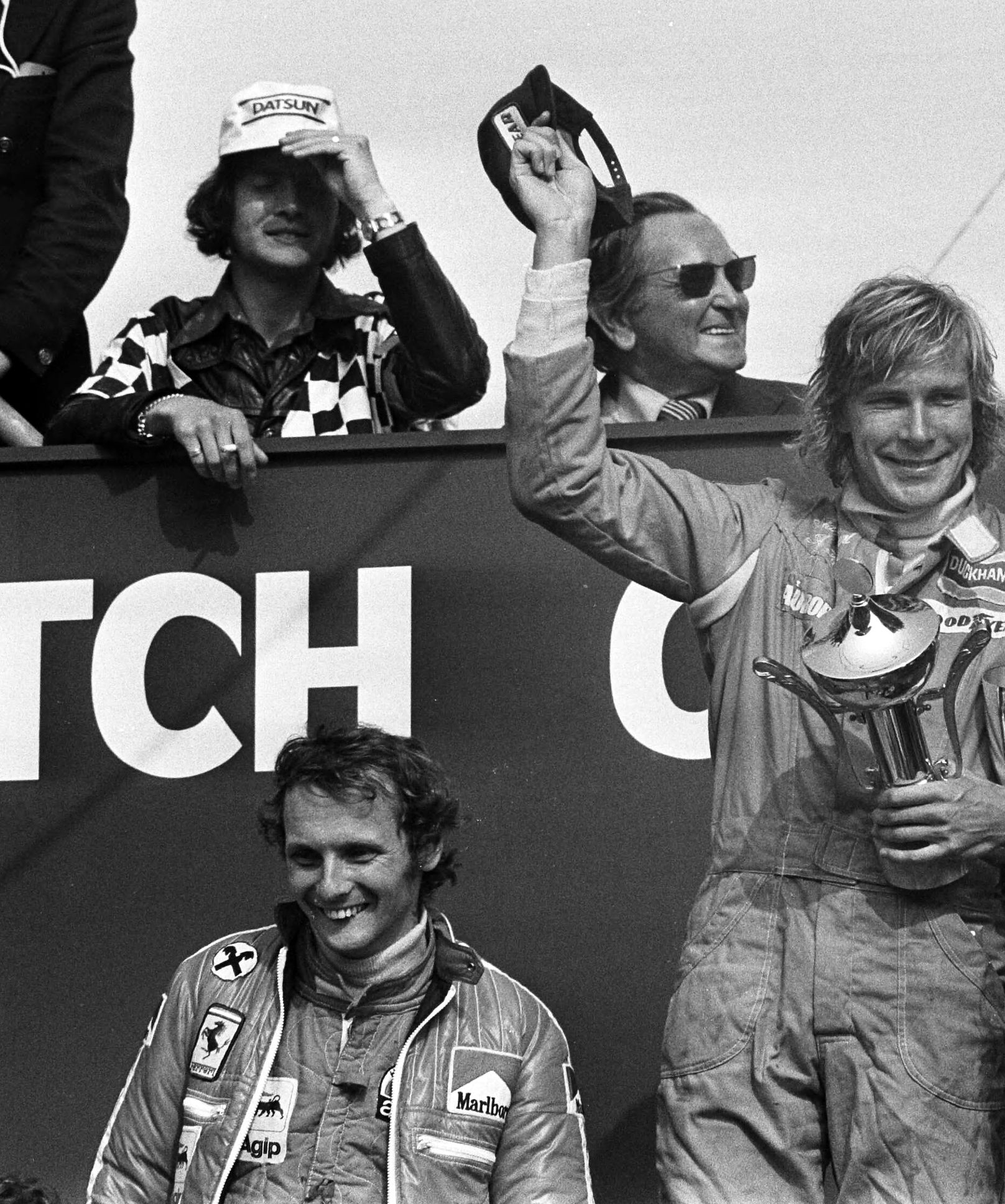 On a podium before a large dark banner with white lettering, a long-haired driver in overalls raises a cap in one hand and holds a small cup in the other; below him another driver in quilted overalls laughs. Behind them two men lean on a rail, one in a checked jacket and a cap.