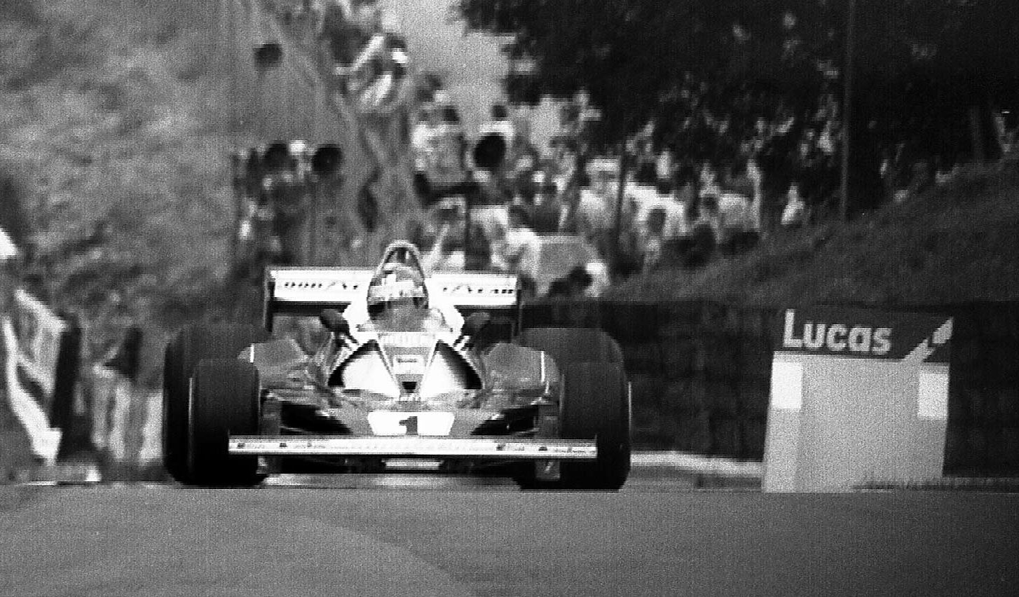 Head-on, a single-seater with a large rear wing and the number 1 on its nose crests a rise towards the camera; spectators crowd a wooded bank behind it, and an advertising board stands at the right.