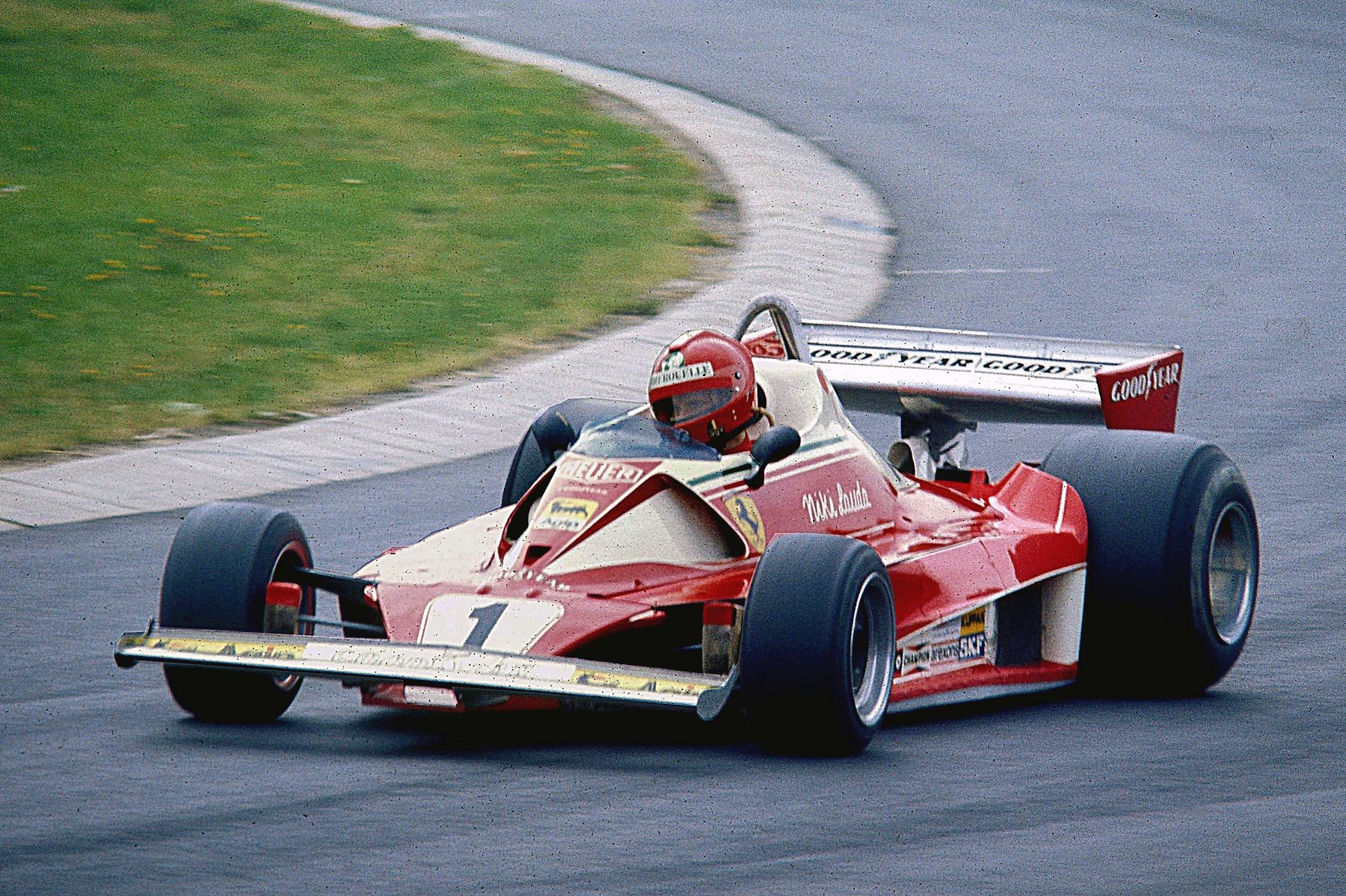 In colour: a red and white single-seater numbered 1, its driver in a red helmet, turns through a bend on grey tarmac beside a grass bank, its tall rear wing lettered with a tyre maker’s name.