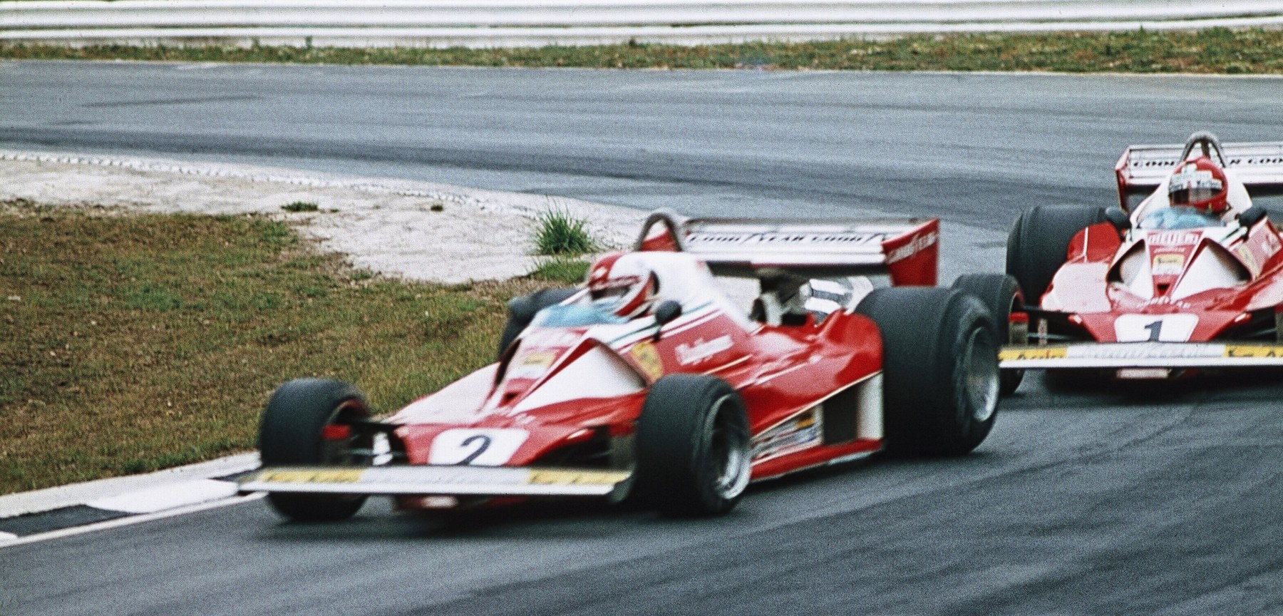 In colour: two red and white racing cars with tall rear wings take a bend one behind the other, No. 2 in front and No. 1 following, on tarmac beside a grass verge and a pale run-off; a barrier runs along the top.