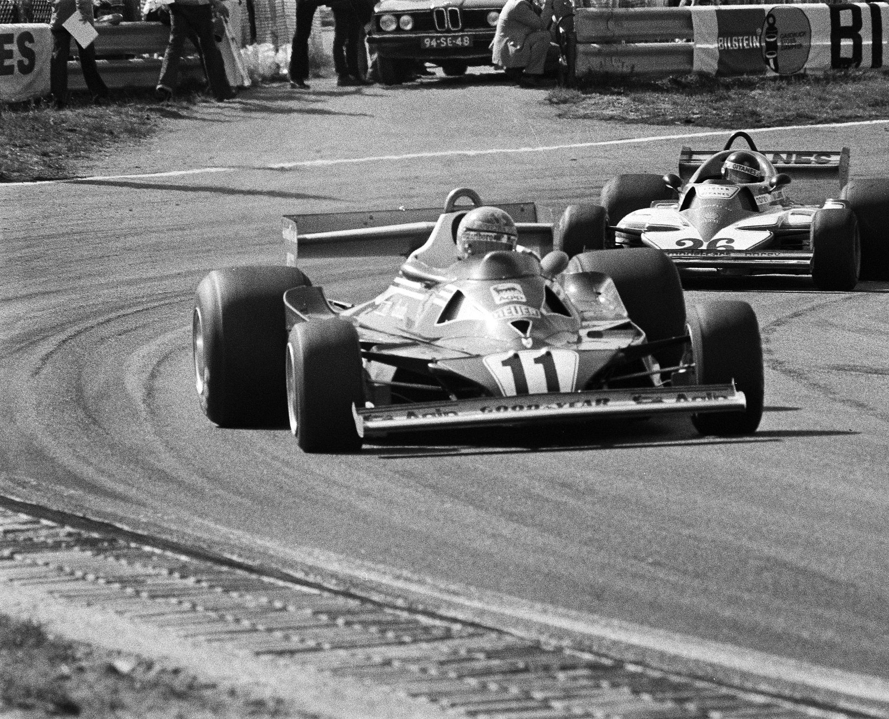 A single-seater numbered 11 leads another car, numbered 26, through a long curve; officials and a parked saloon car stand behind a barrier hung with advertising boards at the top, and a rumble strip runs across the foreground.