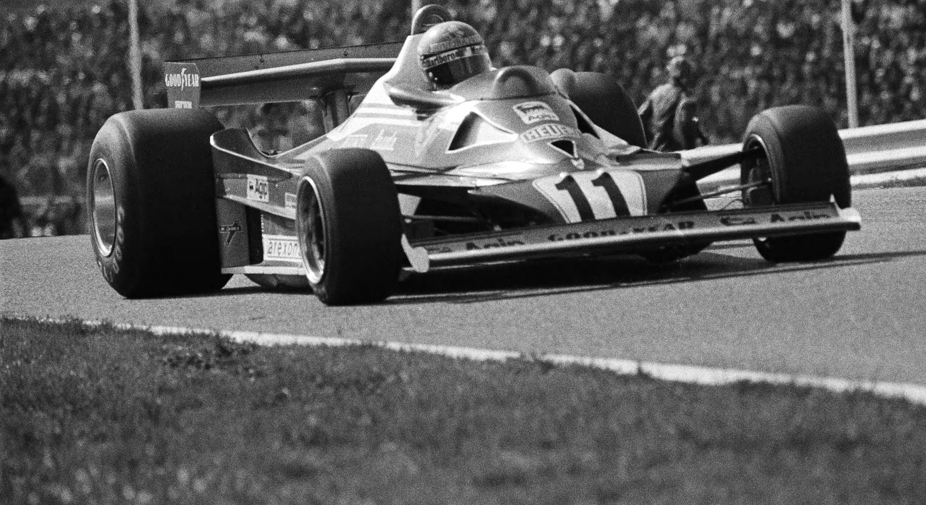 Seen from a low angle, a single-seater, No. 11, with a wide front wing and a tall rear wing leans through a bend towards the camera, the driver’s helmet above the cockpit; a crowded grandstand is blurred behind and grass fills the foreground.