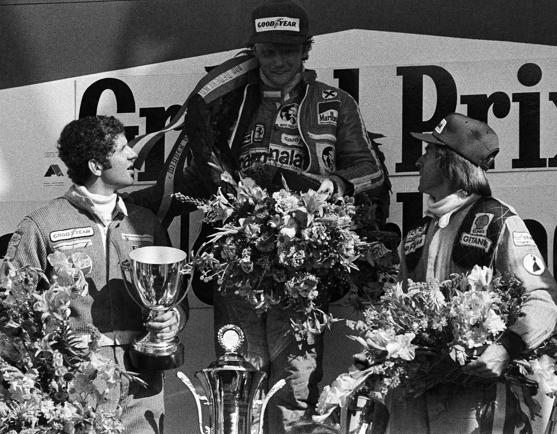 Three drivers on a podium in front of a banner with large lettering; the winner in the centre, in a cap with a ribbon round his neck, holds a great bouquet; the driver at the left holds a cup and looks up at him, and the one at the right, in a cap, turns towards him.