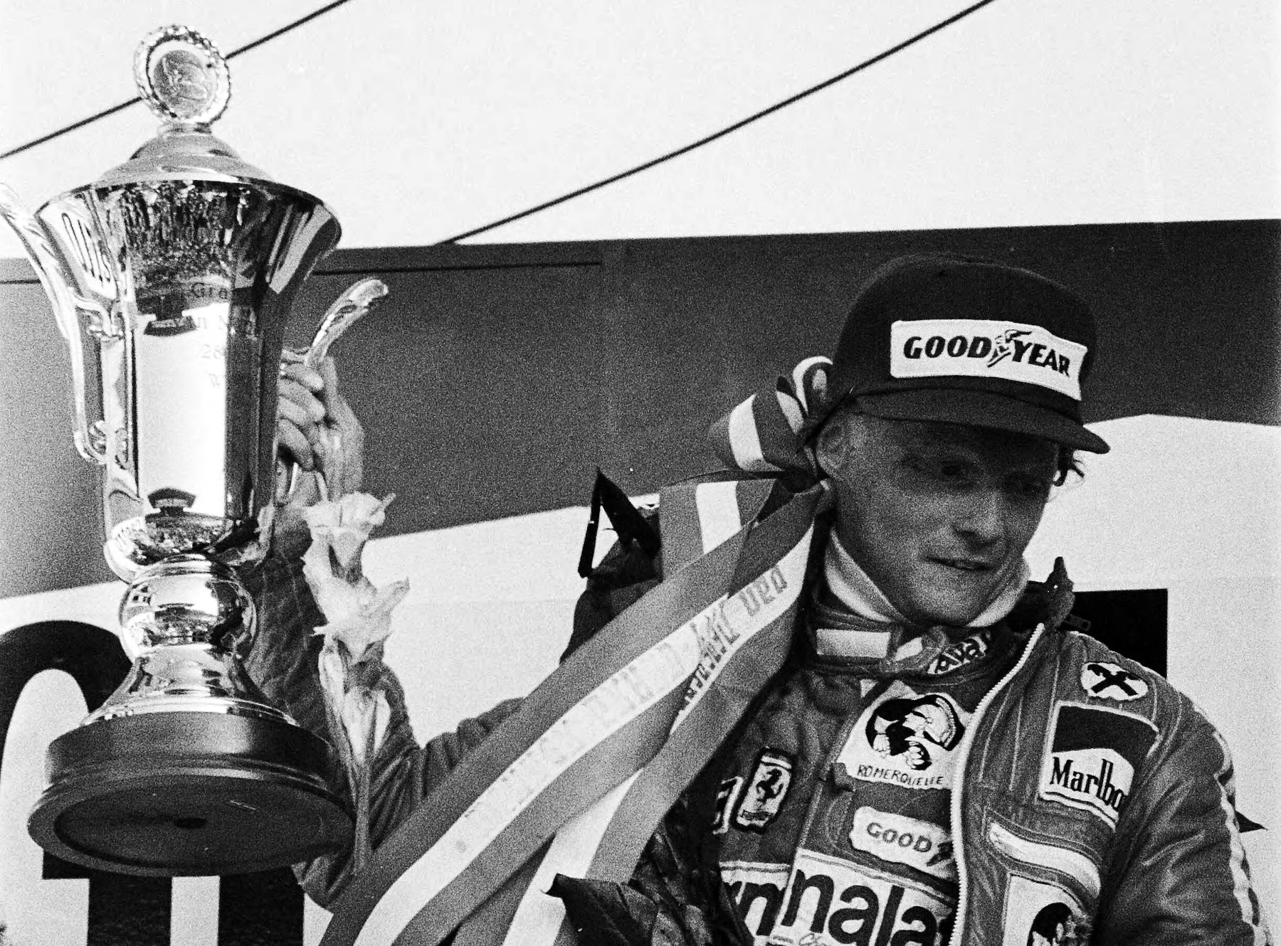 A driver in a cap and racing overalls, a striped ribbon round his neck, holds up a large ornate cup in one hand and looks down, smiling; overhead wires cross the pale sky behind him.