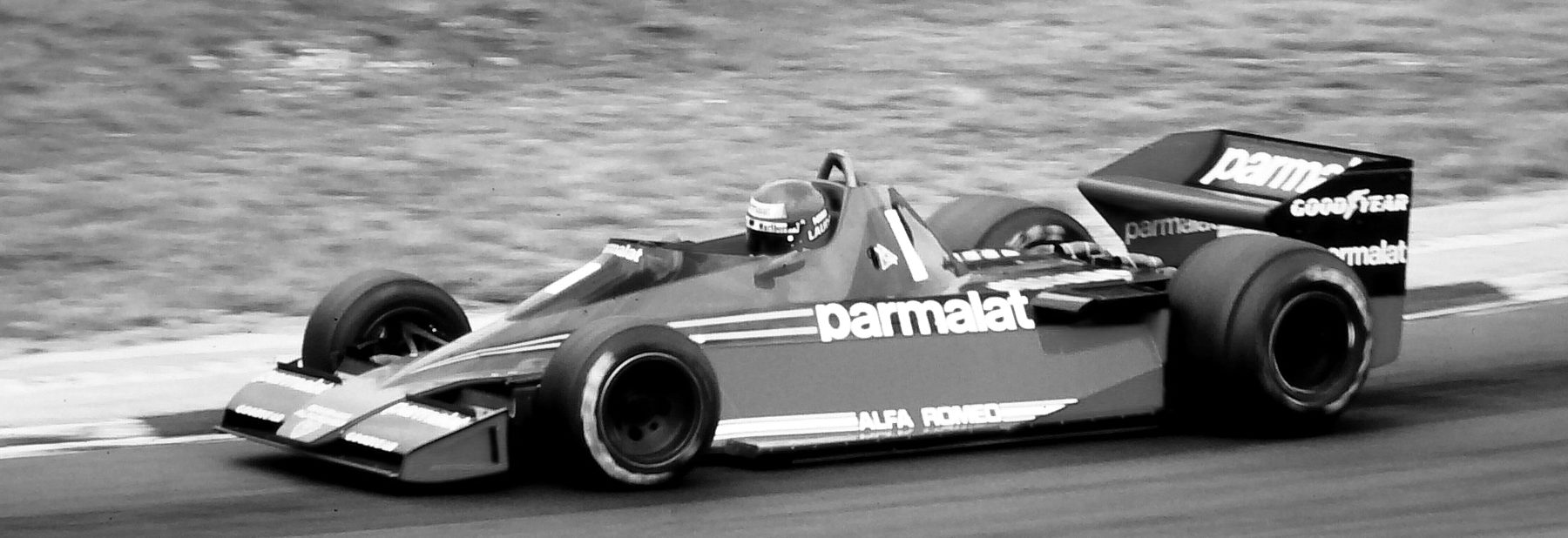 Side view of a single-seater numbered 1, sponsor lettering along its flank and the driver’s helmet above the cockpit, panned so that the grass bank behind it is a blur.