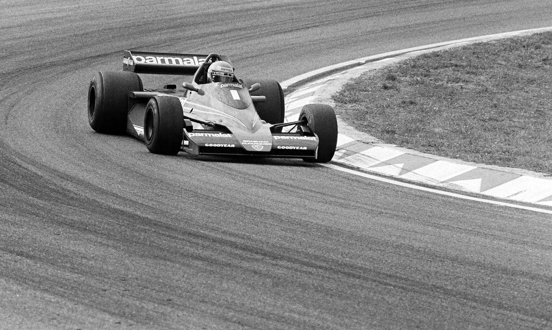 A single-seater numbered 1, seen from the front, leans into a long bend beside a painted kerb, tyre marks curving across the tarmac.