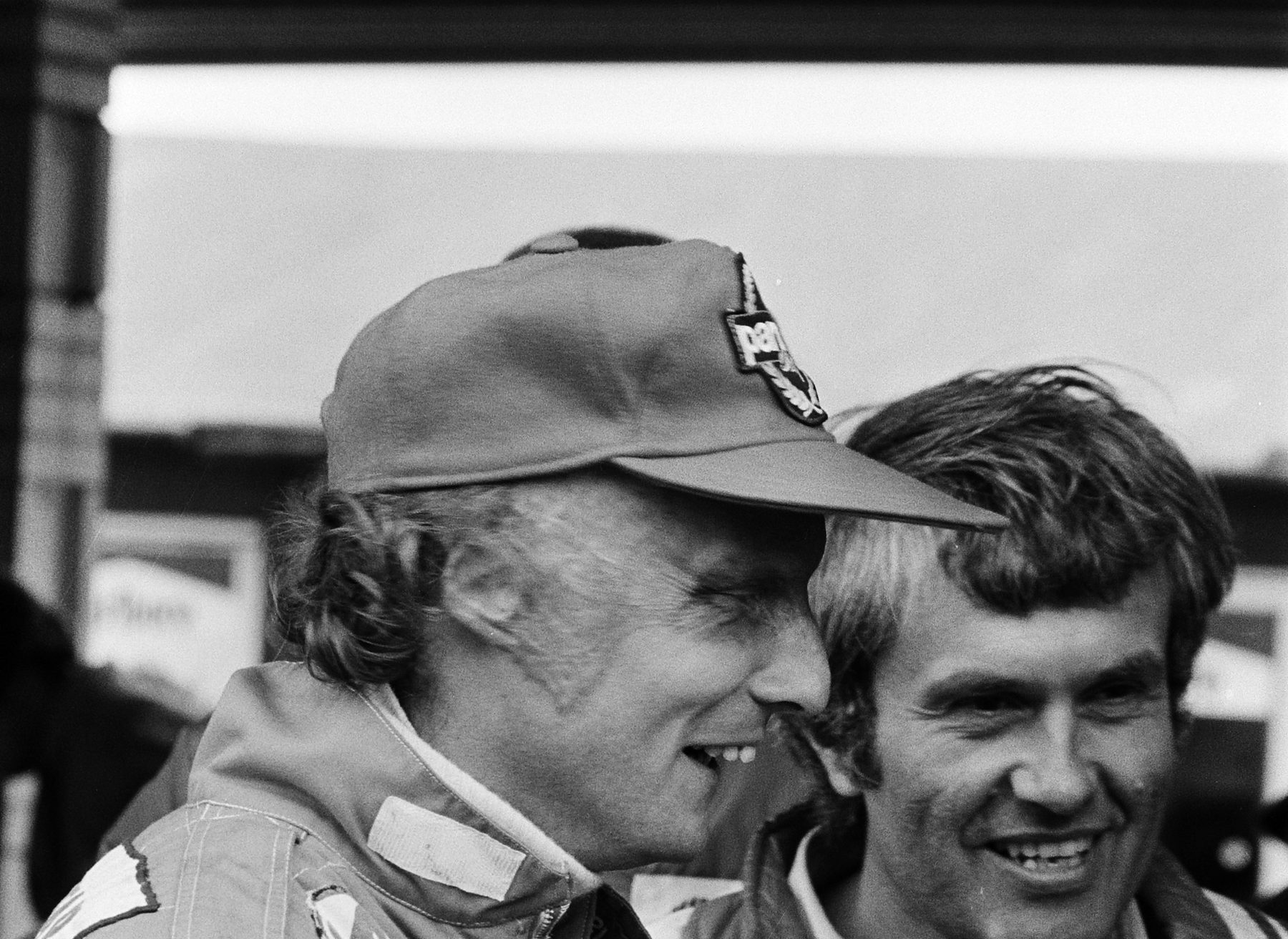 Two men laugh together: on the left, in profile, a man in a peaked cap with a badge and racing overalls; on the right, facing the camera, a man with tousled hair.