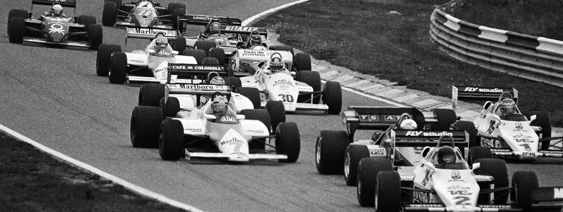A pack of single-seaters crowds through a bend: one numbered 7 leads the left-hand line, a similar car numbered 8 a few places back, with others numbered 30, 26 and 2 around them and a curved barrier behind.