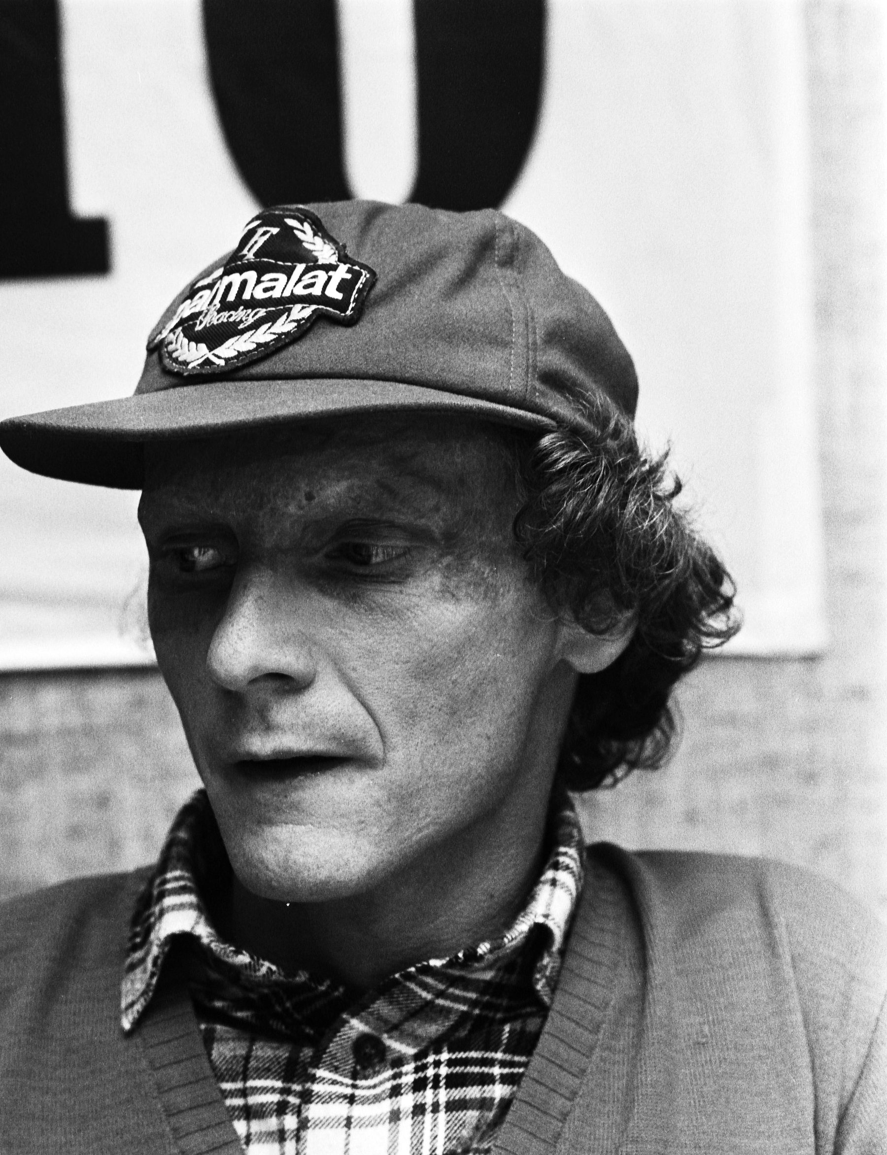 Head and shoulders of a man in a peaked cap with a badge, a check shirt and a V-neck sweater, looking to one side; large dark lettering on a white banner behind him.
