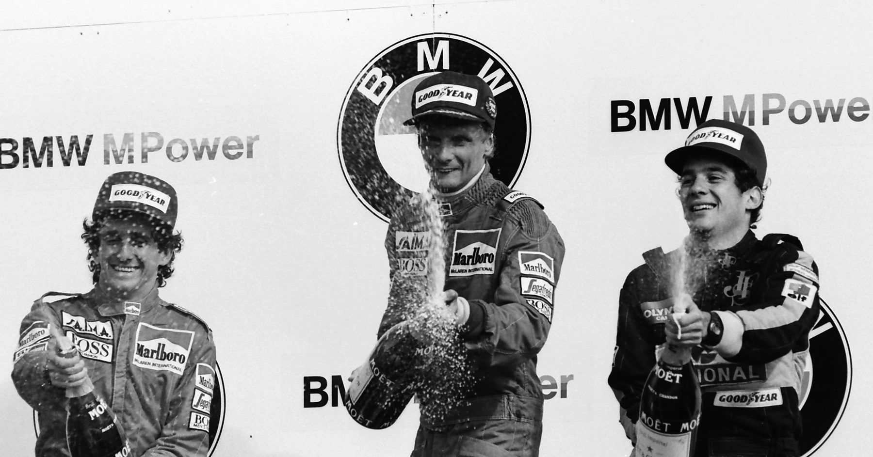 Three drivers in caps on a podium in front of sponsor boards spray champagne: the man in the centre aims his bottle forward, the one on the left laughs, the one on the right grins as his bottle foams.