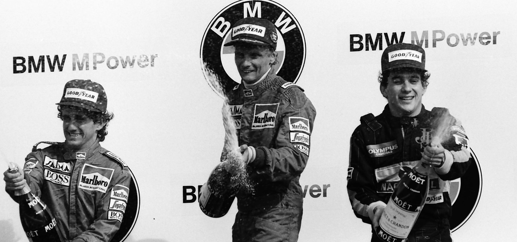 Three drivers in caps and racing overalls stand on a podium in front of sponsor boards, each with a bottle of champagne; the man in the centre sprays his, the two beside him are laughing.