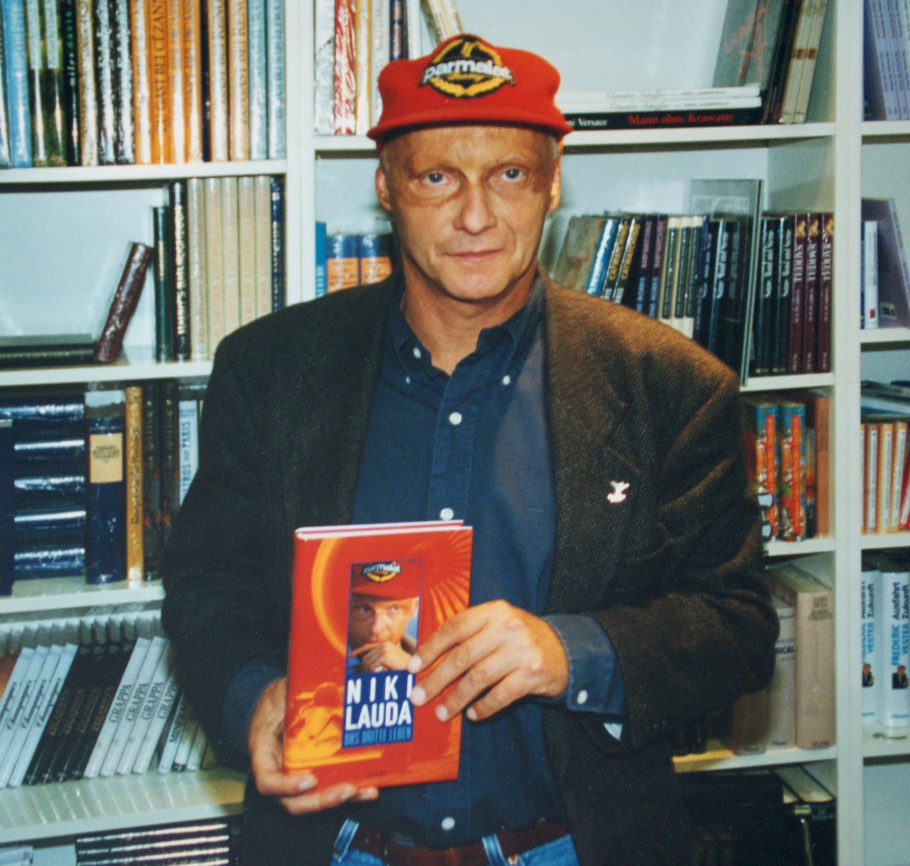 In colour: Lauda in a red sponsor’s cap and a tweed jacket over a dark shirt stands before bookshelves, holding up a hardback with his own face and name on the cover.