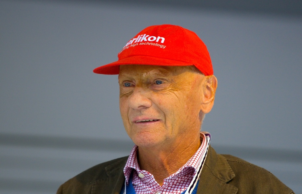 In colour: head and shoulders of Lauda, smiling, in a red sponsor’s cap, a checked shirt and a tweed jacket, against a plain grey wall.